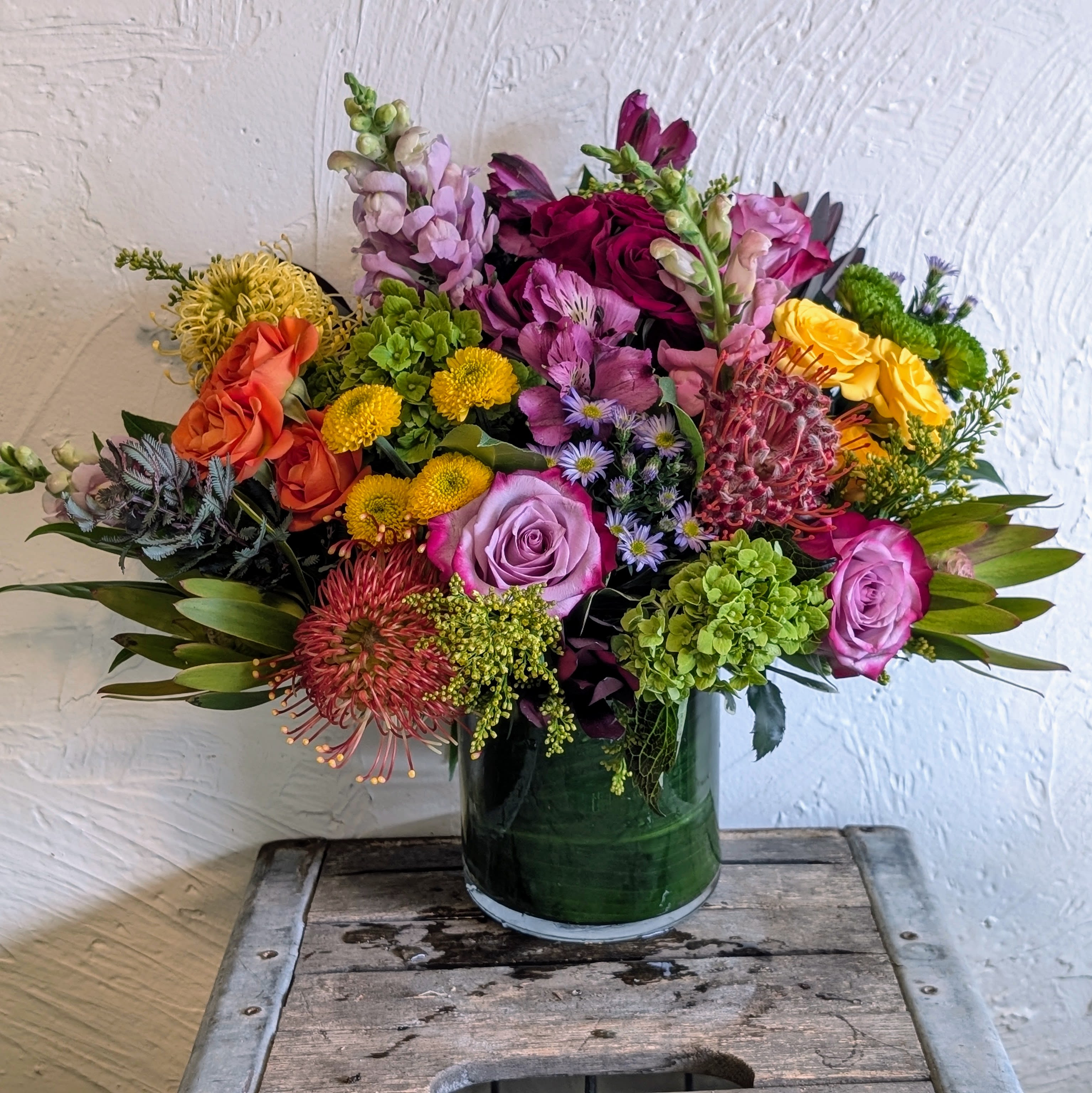 Bloom Boom Pow - A lively, garden-style arrangement bursting with color, perfect as a birthday bouquet or congratulations gift. This flower arrangement blends pink and purple with bright yellow and orange accents to create an immediate, joyful impression - the kind that makes the person you're thinking of feel celebrated the moment it arrives.  Dense and rounded in a dark green glass vase, the composition layers hydrangea clusters, snapdragons, chrysanthemum and spray roses, plus pincushion protea flowers for contrast and texture. The mix of large blooms and smaller filler flowers gives a rich, hand-arranged look with touches of green peeking through; dimensions are compact yet abundant, ideal for tabletops or a reception desk.  Send this to mark a birthday, graduation, or just because - it reads as both festive and thoughtful. Care is simple: keep water fresh and trim stems every few days to prolong bloom life; the sturdy green vase arrives ready to display.