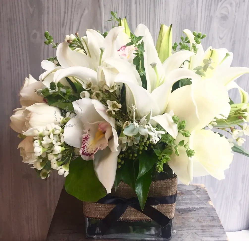 White lilies and orchids arranged in a square glass vase with a ribbon wrap.