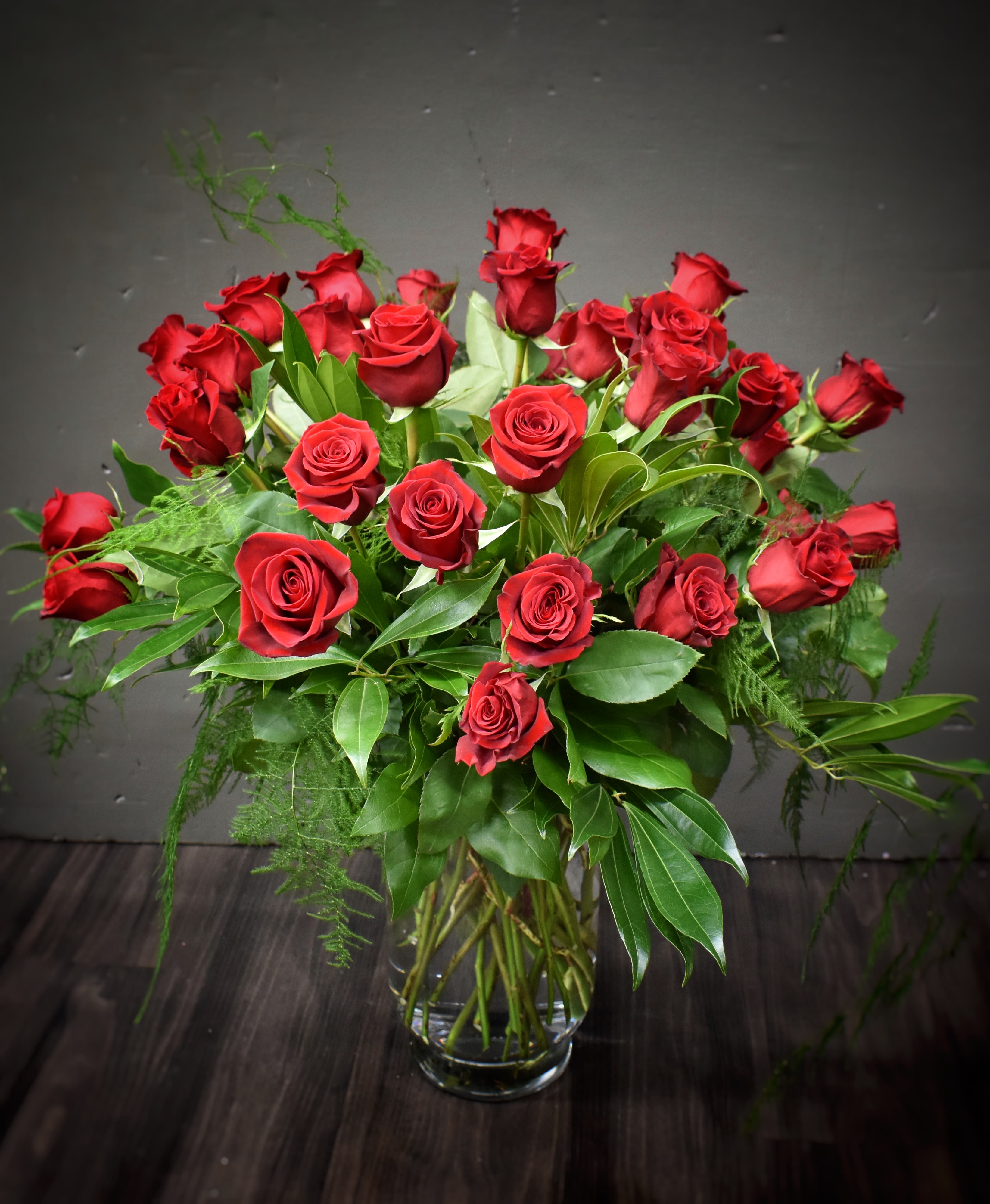 Tall arrangement of long-stemmed red roses with lush foliage in a clear glass vase