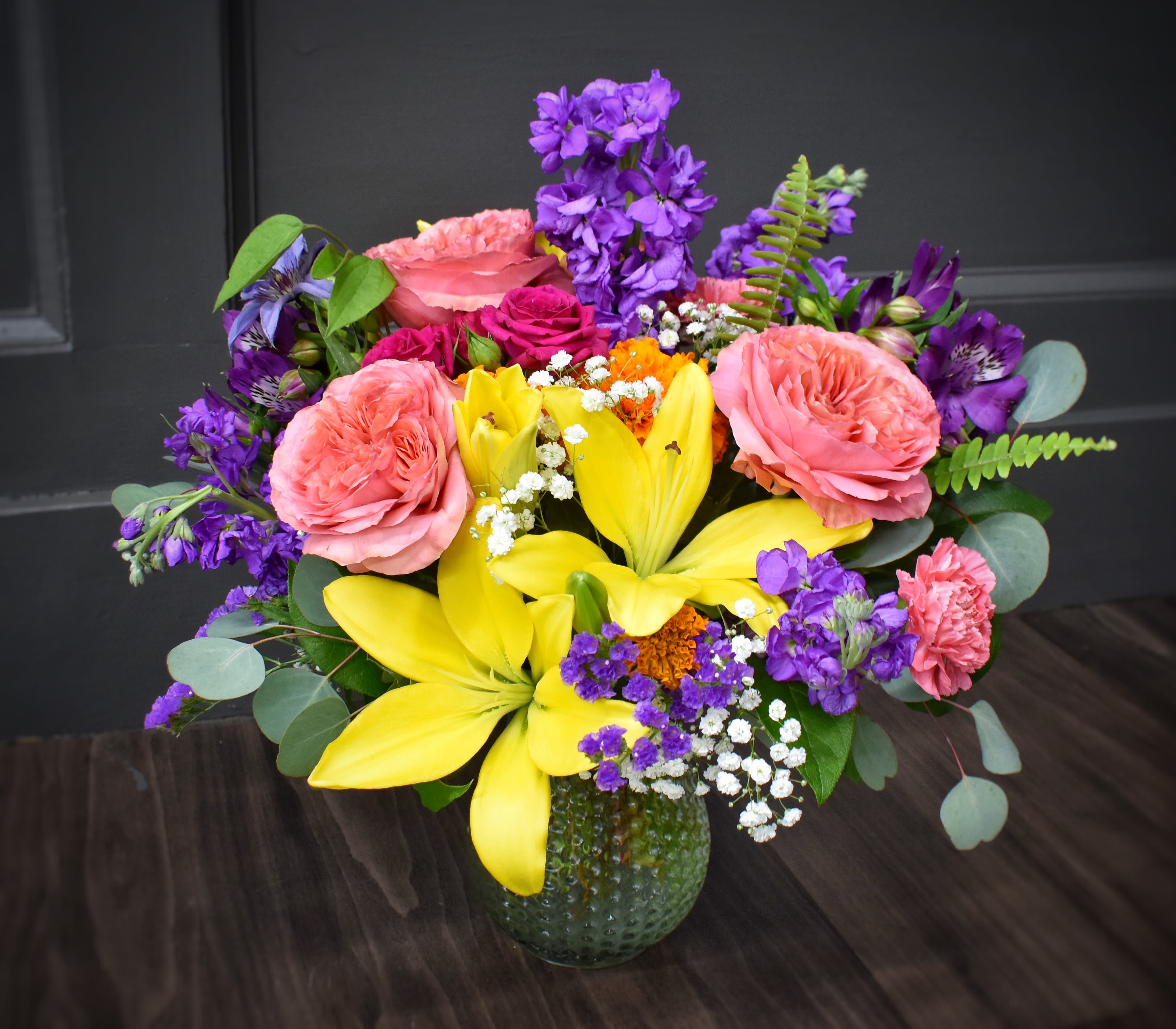 Color Splash! - PREMIUM - Bright yellow lilies burst amid pink roses and ruffled carnations in a jubilant mixed-color bouquet made for birthdays, thank-you gestures, or any cheerful spring celebration. This lively flower arrangement puts the occasion front and center: the person you're surprising will feel noticed and uplifted by the bold contrast of sunny yellows, soft pinks, and pops of purple.  The arrangement pairs large lilies, garden-style roses, carnations, and clusters of purple stock with delicate white filler blooms for texture and scent. It's arranged in a green textured glass vase that gives a modern, slightly retro anchor to the rounded, full composition-easy to set on a table or gift as a ready-made centerpiece. Sized to feel generous without overwhelming a room, it's a fresh, celebratory choice that looks great from every angle; simple care-fresh water and a cool spot-keeps it vibrant for days.