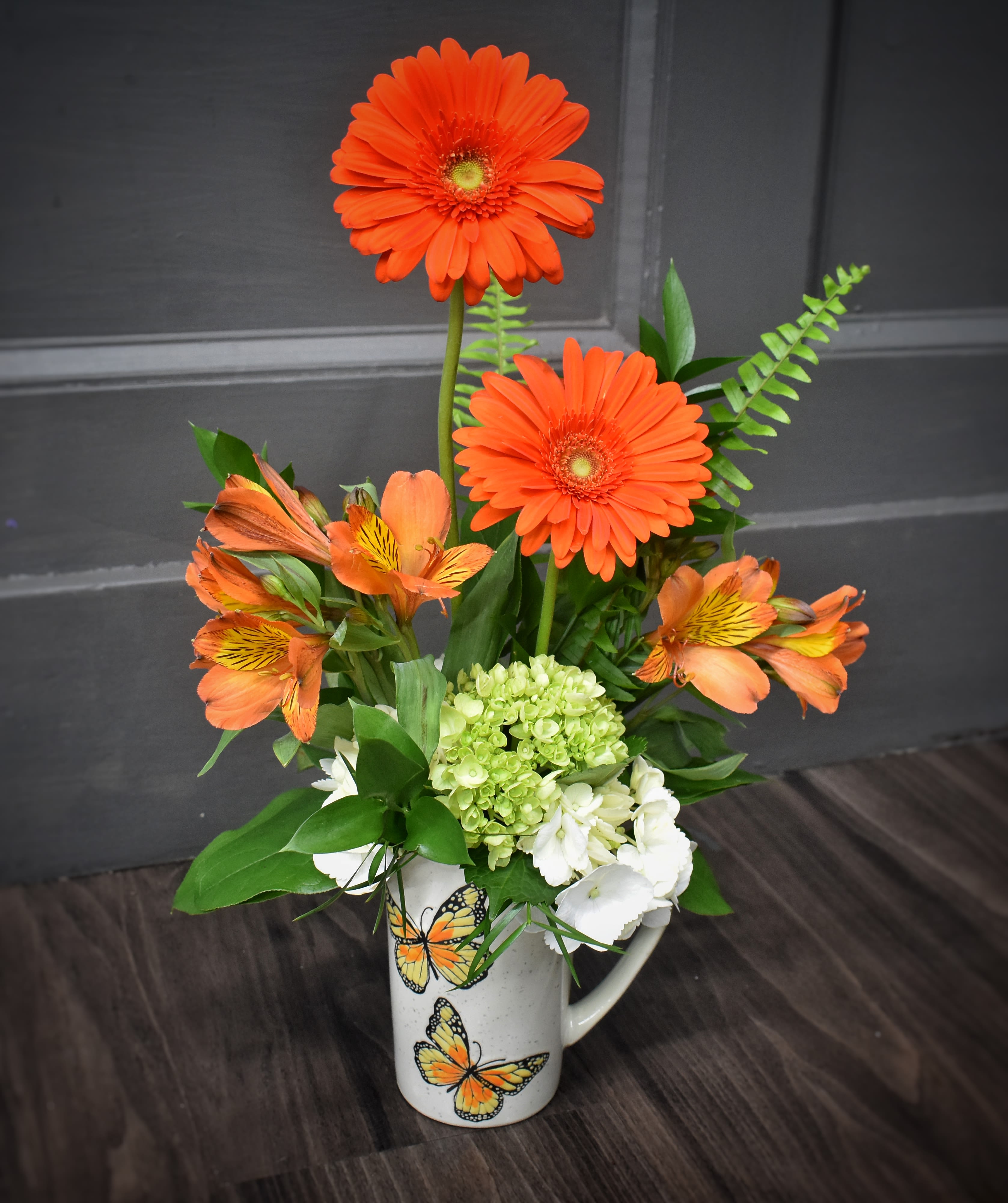Flutterby Blooms - Bright orange gerbera daisies pop above a cheerful cluster of orange alstroemeria, pale green hydrangea, and crisp white accents in this playful flower arrangement-perfect as a birthday bouquet, thank-you gift, or just-because pick-me-up. The butterfly-adorned ceramic mug gives it a whimsical, everyday charm that reads like a sunny surprise for the person you're thinking of.   Flowers are arranged  inside a white ceramic mug embossed with butterflies; the set arrives with a  bag of pimento cheese popcorn for an extra bit of delight. It's the kind of casual, uplifting gift that makes someone feel seen-easy to display on a desk or kitchen counter and ready to brighten the day.