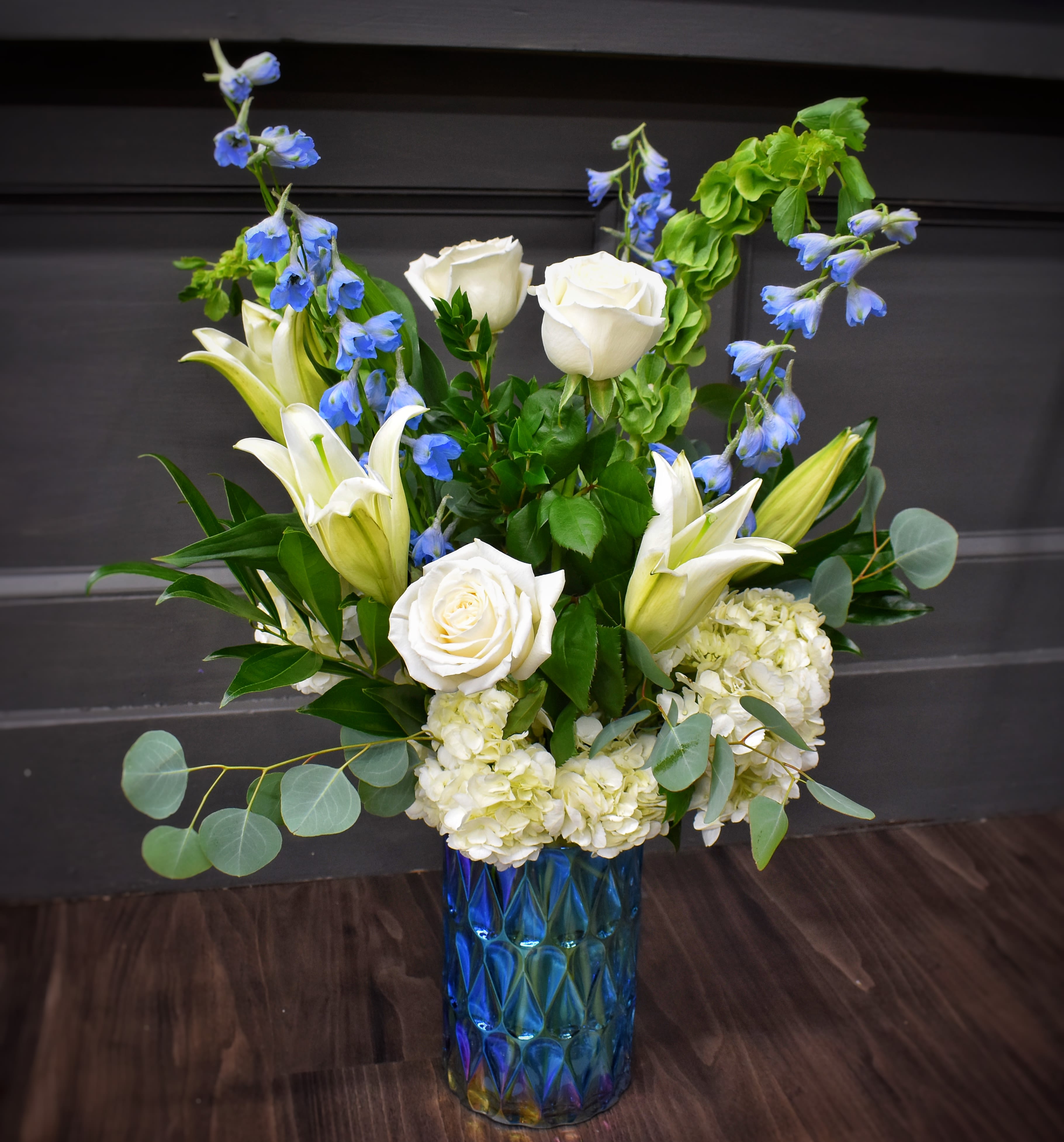 Blue Skies - Blue Skies greets the recipient with a calm, airy palette of white and blue that feels both comforting and celebratory-perfect as a sympathy arrangement, a thoughtful get-well gift, or a refined thank-you bouquet. The vertical, slightly loose design lets tall blue delphinium spikes arch above clusters of white roses and lilies, creating an uplifting silhouette that reads as serene and elegant for everyday gifting or more formal moments.  White hydrangea adds soft, rounded volume while eucalyptus-like green foliage softens the edges and gives the bouquet a fresh, garden-fresh texture. Arranged in a tall blue glass vase, the jewel-toned base grounds the composition and lets the cool whites and blues pop; the layered look draws the eye from the rounded focal blooms up through the airy blue accents.  