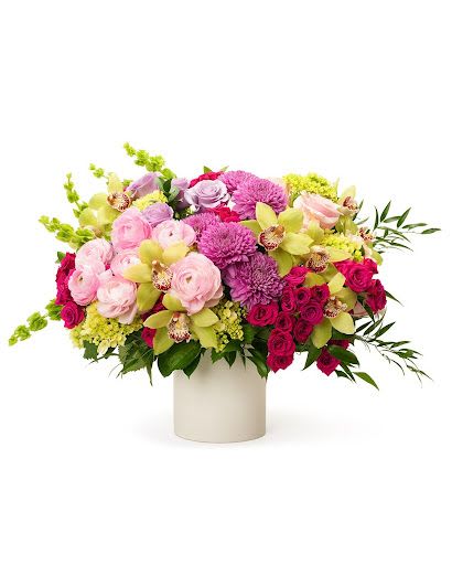 Low arrangement of pink roses, purple mums, and green orchids in a white cylinder vase