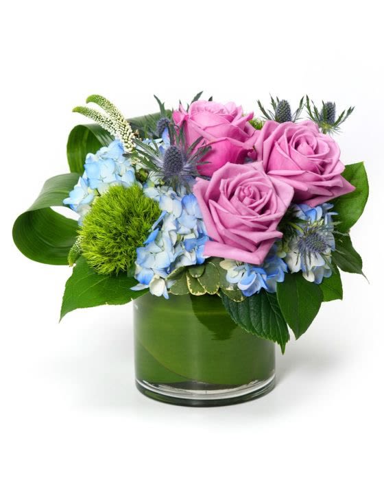 Short arrangement of pink roses, blue hydrangea, and green dianthus in a leaf-lined glass vase.