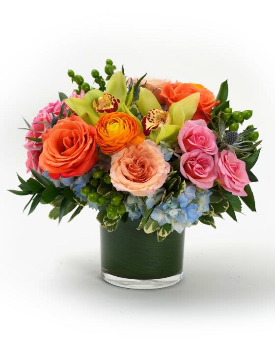 Low glass vase of orange and pink roses with green orchids, orange ranunculus, and blue hydrangeas