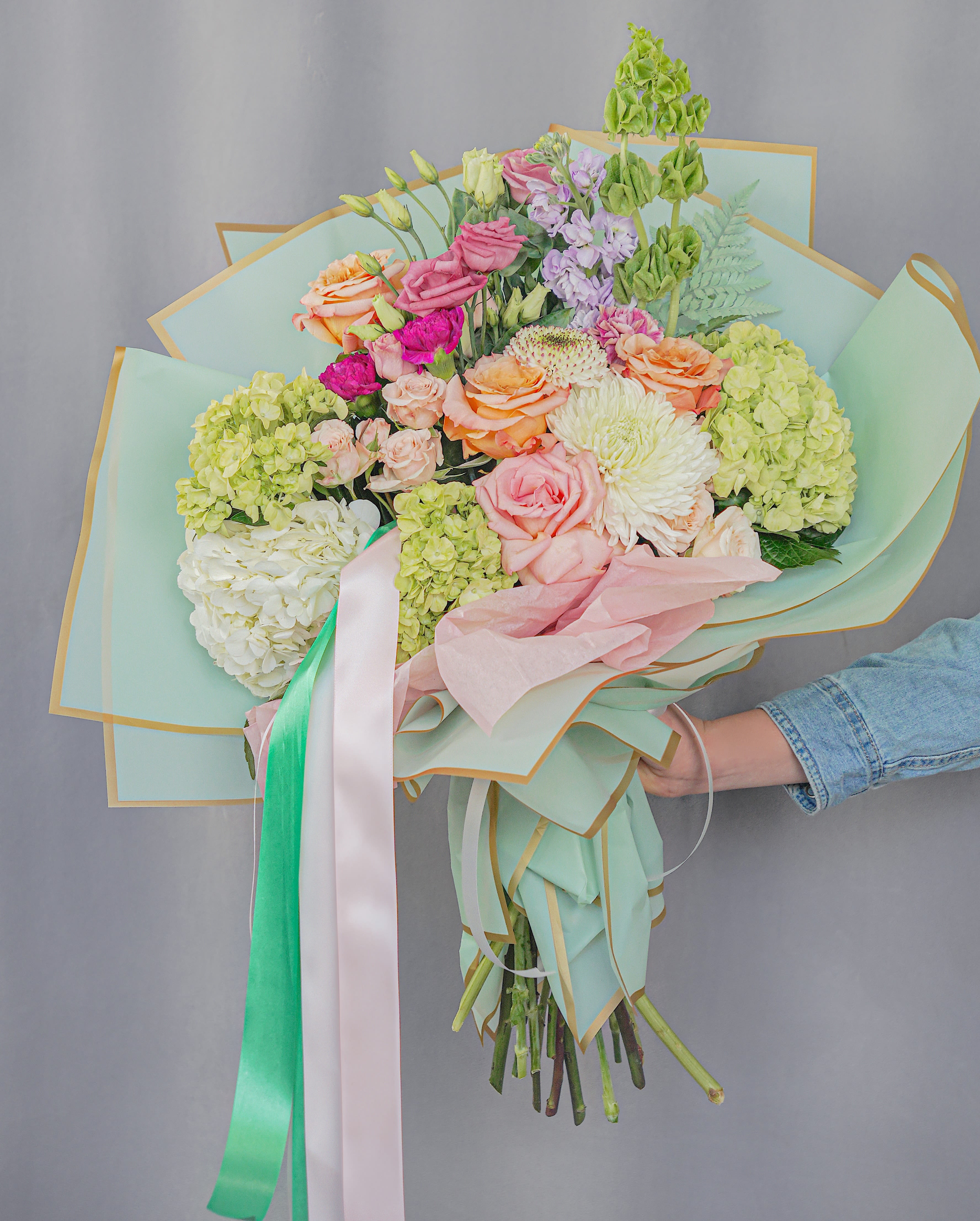 Handheld bouquet of pastel roses, hydrangeas, and chrysanthemums wrapped in mint paper