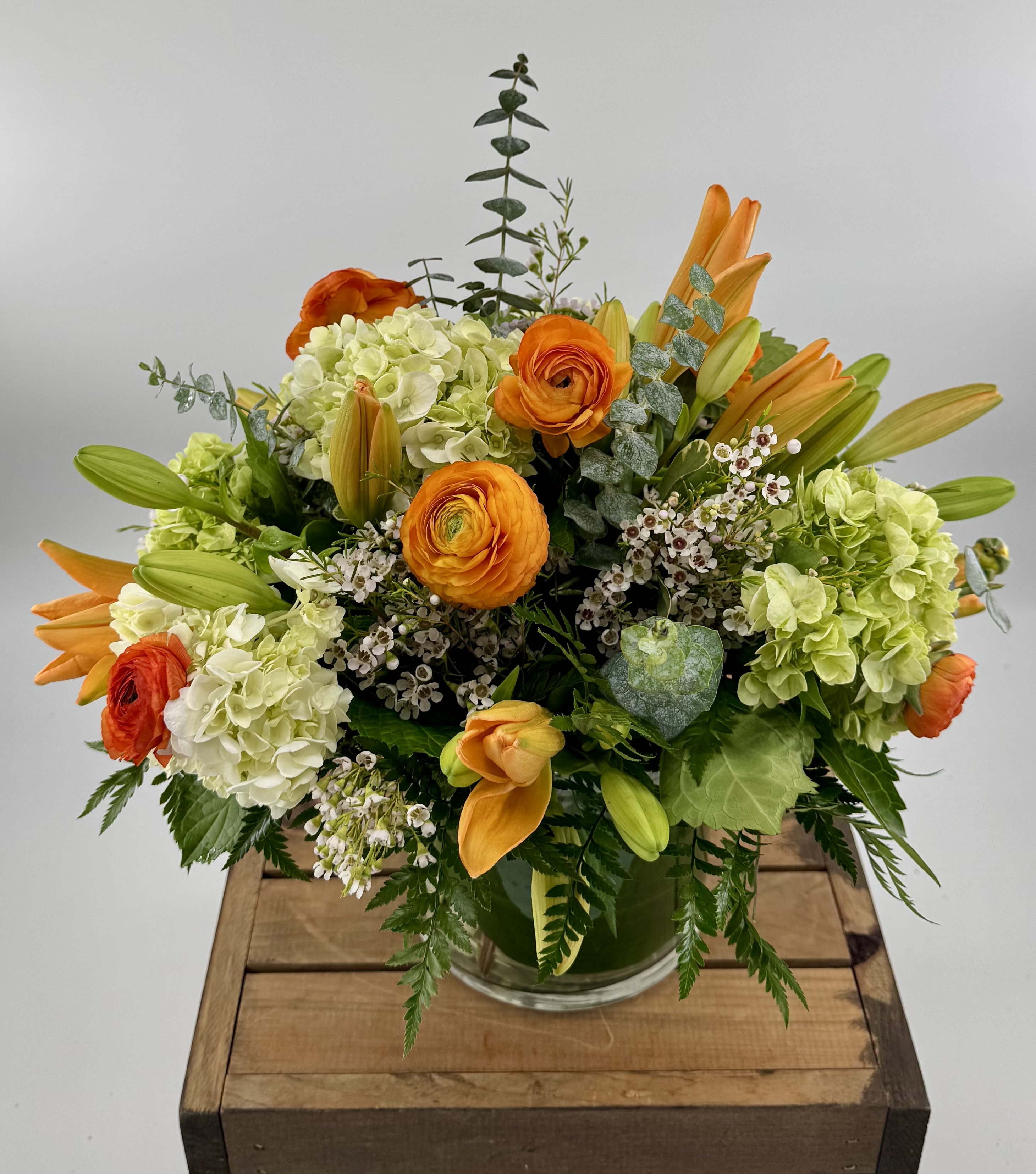 Love Me at Sunset - This luxurious modern arrangement features vibrant dark orange ranunculus, bright orange Asiatic lilies, and delicate wax flowers, expertly designed in a sleek 6x6" glass cylinder vase—perfect for adding a sophisticated pop of color to any space. 