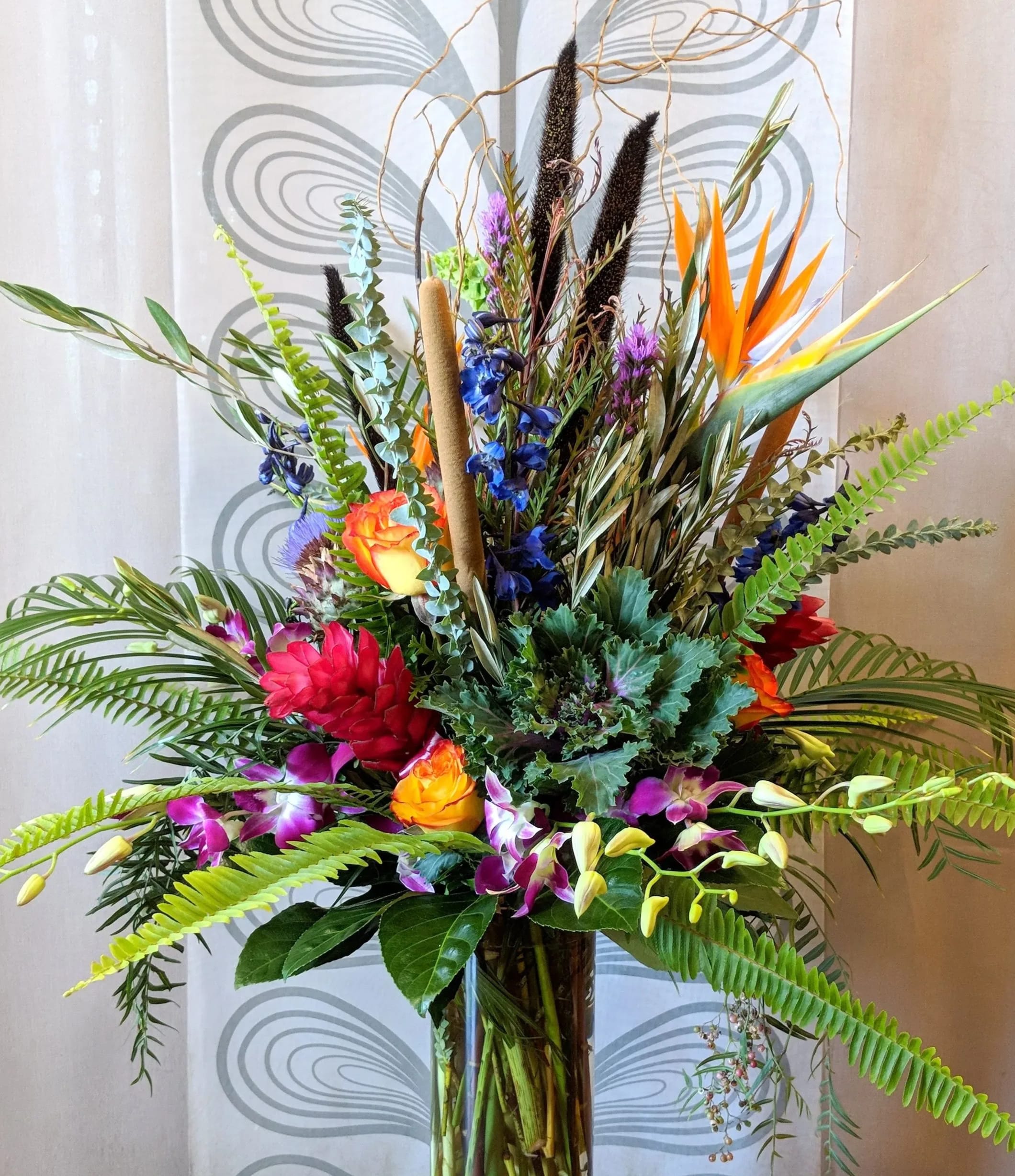 Luscious - Introducing a tall, modern, and show-stopping tropical vase arrangement that will transport you to a vibrant and exotic paradise. This arrangement features a stunning combination of birds of paradise, purple dendrobium orchids, blue delphinium, red ginger, yellow roses, kale, bells of Ireland, and is accented with tropical foliage and curly willow.