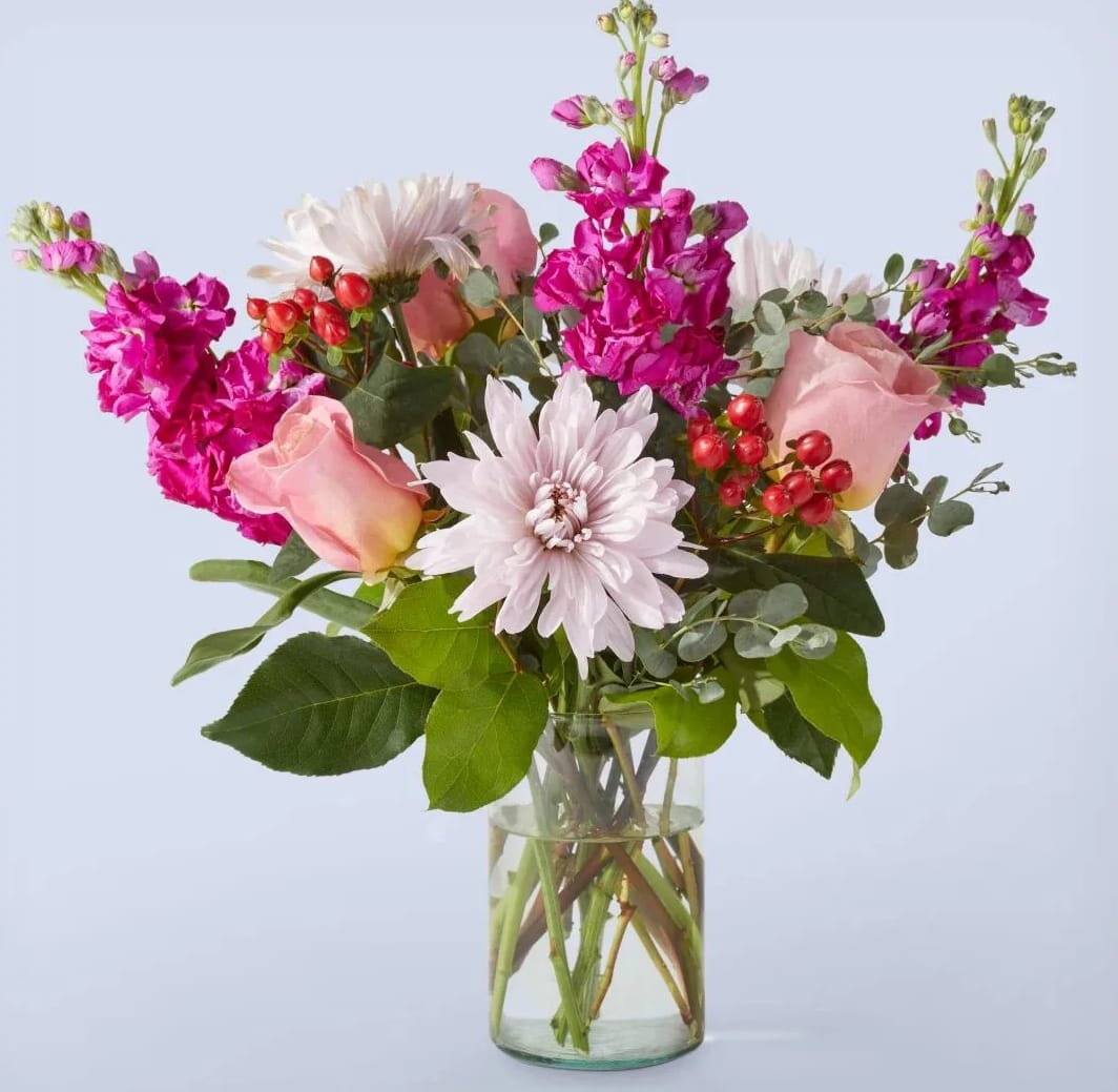 Sunday Morning - Start your day off right with the "Sunday Morning" arrangement, a stunning bouquet that captures the essence of warmth, joy, and tranquility. This beautiful arrangement features a vibrant assortment of pink roses, hot pink stock, purple Fuji mums, and red hypericum berries, expertly arranged in a glass vase with lush greenery.