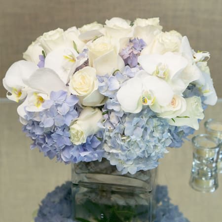 Blue Clouds - Indulge in the serene beauty of our exquisite floral arrangement, featuring the captivating combination of blue hydrangeas, white roses, and white orchids. Expertly arranged in a leaf-lined glass cylinder vase, this ensemble exudes elegance and tranquility.