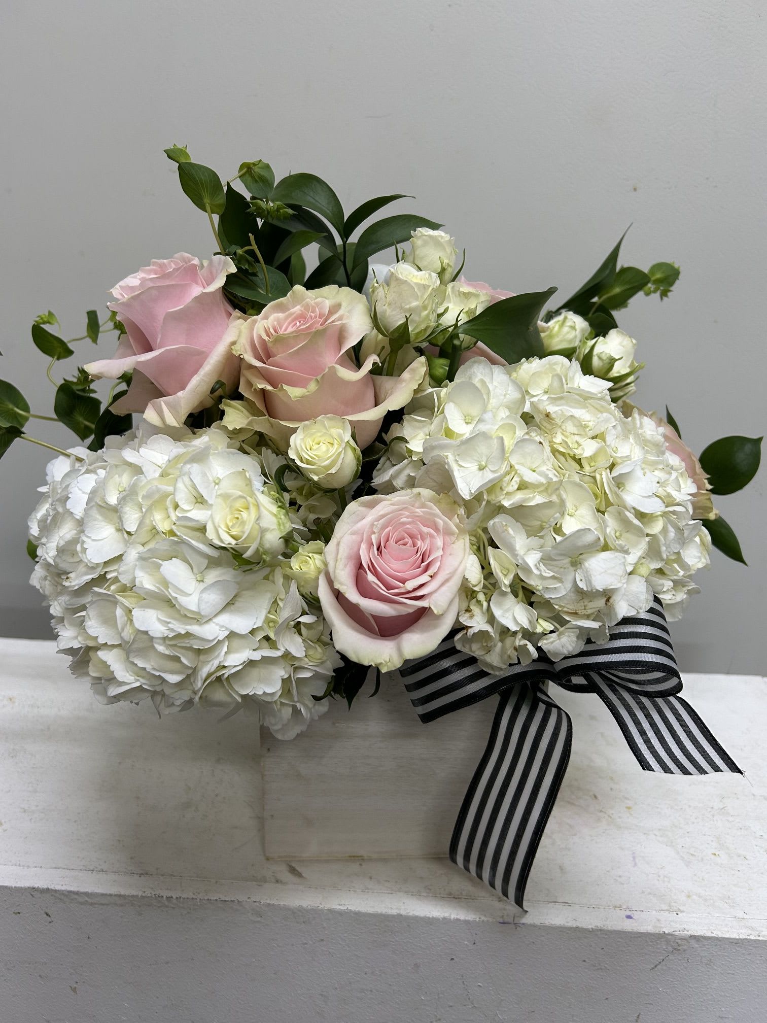 Boxed - Pale pink roses nestled into billowy white hydrangeas create a soft, romantic statement in this compact flower arrangement - perfect as a birthday bouquet, anniversary gift, or a thoughtful way to say thank you. The rounded, close-set presentation focuses every glance on the delicate blooms so the person you're thinking of will immediately feel celebrated and cherished.  Dark green foliage rises behind the flowers to give contrast and structure, while the blooms sit in a clean white box tied with a black-and-white striped ribbon for a polished, gift-ready finish. The combination of roses and hydrangeas in white, pink and green reads refined and modern; its tidy size and box base make it easy to display on a desk, bedside table, or dining table.  Send this when you want to offer quiet romance or sincere appreciation - the kind of arrangement that reads both intimate and elegant. Care: keep blooms in a cool spot away from direct sunlight and refresh the water as needed to extend vase life; the boxed presentation makes it simple to place where it will be most admired.