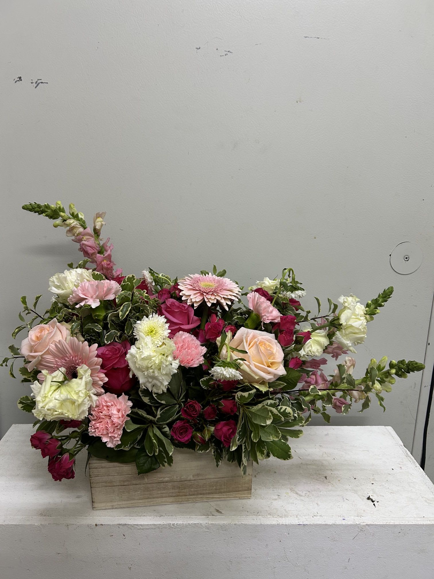 Chisolm - Lush blush roses and bold pink gerbera daisies spill from a rustic wooden box in this garden-style flower arrangement-perfect as a birthday bouquet, anniversary surprise, or any celebration that calls for bright, romantic cheer. The design reads feminine and joyful at a glance, so the person you're thinking of will feel seen and celebrated the moment it arrives.  This low, rectangular arrangement layers roses, gerbera daisies, carnations and clustered mixed blooms in shades of pink, white, and deep magenta, with upright snapdragon spikes adding vertical movement on both sides. The wooden box gives a warm, casual contrast to the soft petals and creates a full, slightly asymmetrical silhouette that looks freshly gathered from a garden.  Sized to sit beautifully on a dining table or console, Snappy makes an immediate impression without needing a tall vase-no-fuss styling that keeps the focus on the flowers. For longest enjoyment, keep blooms in a cool spot away from direct sun and replace water as needed; it's an effortless, heartfelt way to mark a special moment.