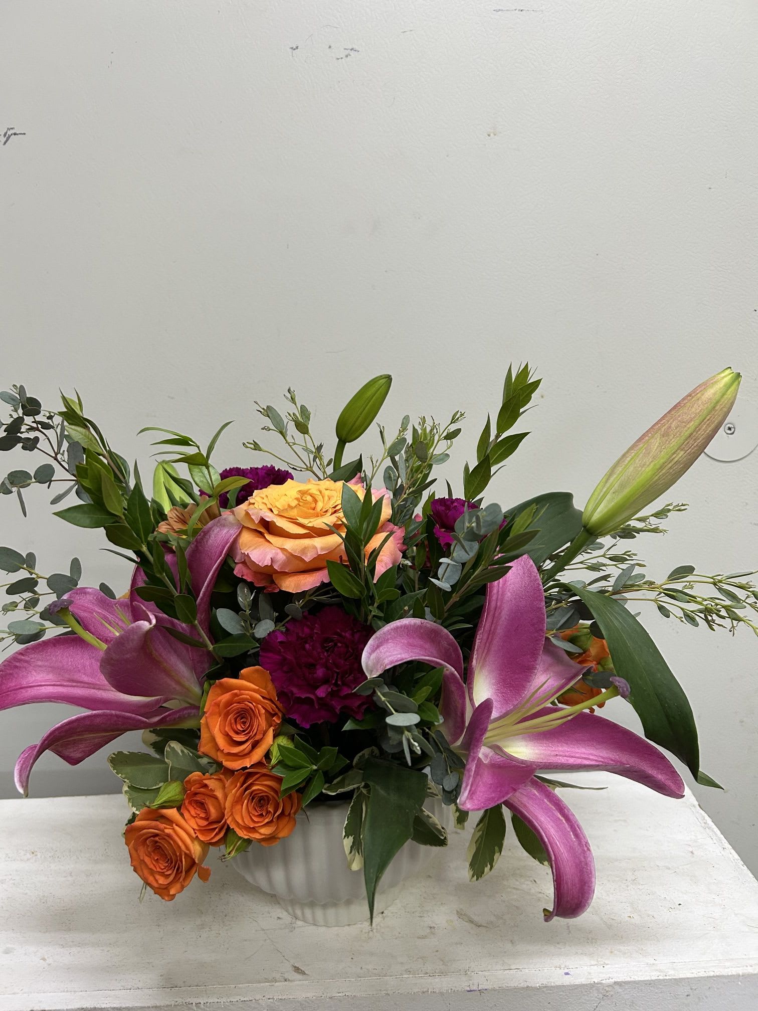 Lily Bloom - Vivid orange roses and large pink‑purple lilies combine with deep magenta carnations in a lush, garden‑style flower arrangement perfect for a birthday bouquet or cheerful thank‑you gift. The energetic contrast between warm orange blooms and cool pink‑purple lilies creates an immediate sense of celebration-she'll feel genuinely seen the moment this arrives.  Arranged low and full with stems and foliage extending outward, the composition reads layered and alive from every angle. Delivered in a simple white ceramic vase, the presentation stays clean and modern while letting the colors take center stage. Ideal for everyday gifting or brightening someone's day; enjoy the arrangement on a table or console where its color and texture can be admired up close.