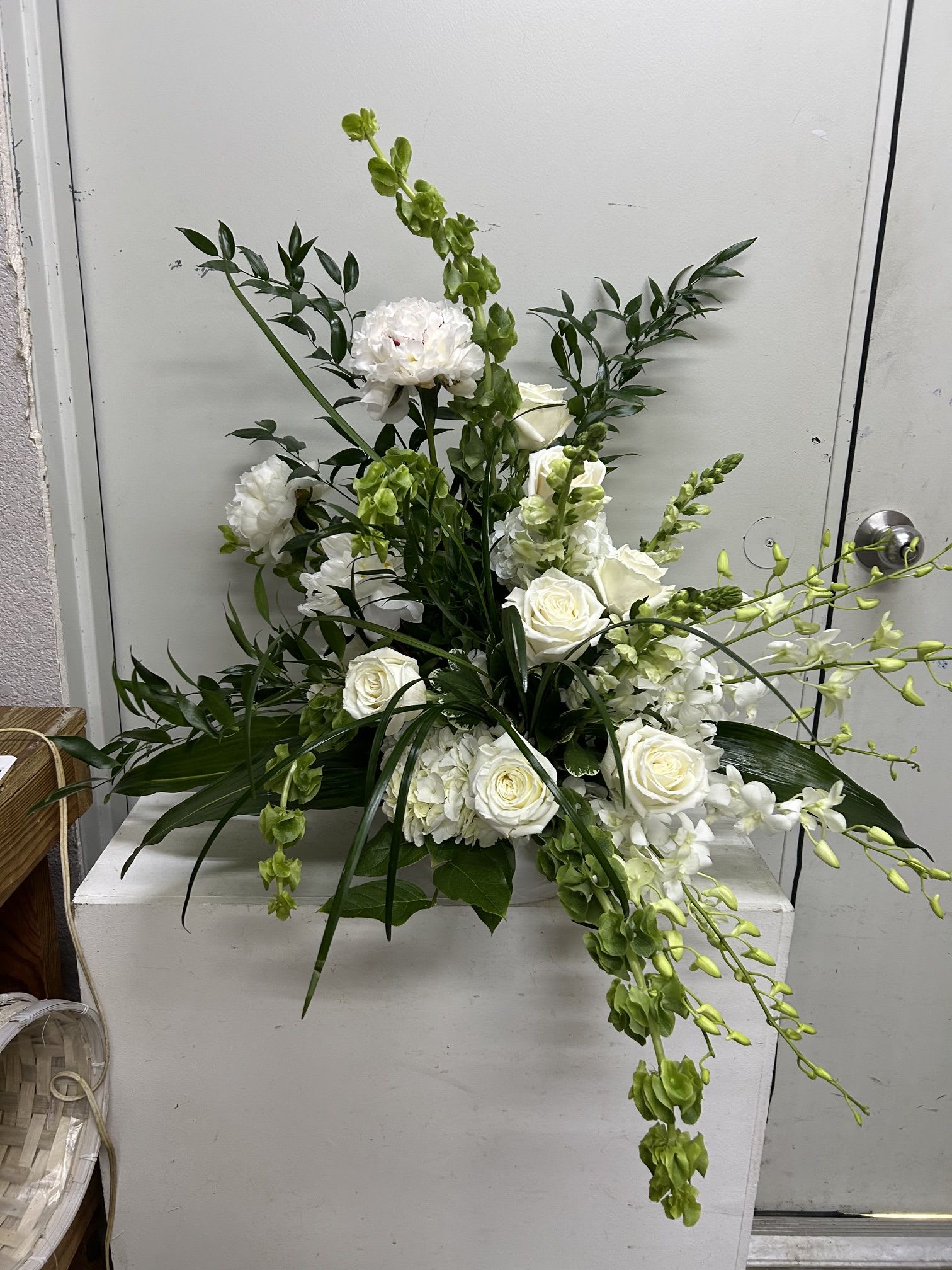 Marie - White roses and clustered white hydrangeas form a calm, refined focal point in this sympathy-style flower arrangement, designed to convey sincere, understated comfort. The tall green snapdragon spikes and dark glossy foliage create an airy, asymmetrical silhouette with cascading stems that give the design gentle movement-perfect for a memorial, funeral service, or any moment when you want the person you're thinking of to feel honored and held.  Crisp white roses and voluminous hydrangeas are paired with vertical snapdragons and deep green leaves to balance softness and structure; the palette of white and green keeps the mood peaceful and formal without feeling heavy. The arrangement's elongated, cascading shape makes it suitable for placement on an altar, beside a casket, or on a mantel where its graceful lines can be seen from several angles.  Choose this arrangement to express sympathy, respect, or quiet remembrance-the kind of floral tribute that makes a grieving family feel comforted and seen. Standard sizing and care are handled by our florist so blooms arrive fresh and properly hydrated; for delivery timing or to add a personal ribbon or message, please contact us when ordering.