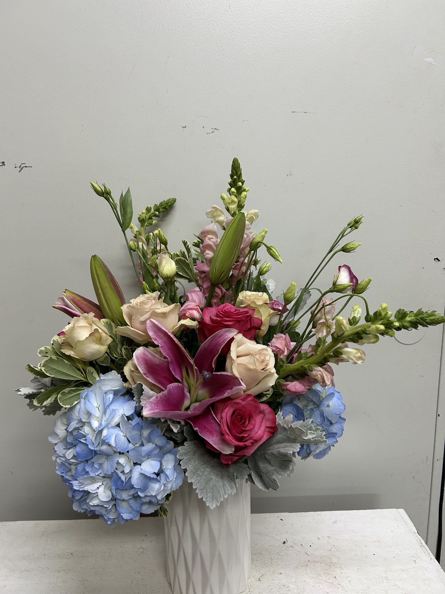 Mix It Up - A lively, romantic flower arrangement that feels like a joyful celebration in bloom - perfect as a birthday bouquet, anniversary surprise, or a cheerful thank-you gift. This Mix It Up flower arrangement places pink and pale blush roses alongside saturated magenta lilies so the person you're thinking of will immediately feel adored; the overall look reads as festive yet thoughtful for everyday moments that deserve attention.  Bright blue hydrangea clusters anchor the lower half with a full, rounded shape while upright snapdragon spikes and airy lisianthus create height and graceful movement. Accents of silver-gray foliage and fresh green stems add cool contrast against the pinks and blues, all arranged in a tall white ceramic vase that suits a dining table, entryway console, or bedside. The combination of textures - velvety roses, ruffled hydrangeas, trumpet lilies, and delicate lisianthus - gives long-lasting visual interest.  Send this to someone who should feel genuinely celebrated: a close friend marking a milestone, a partner on an anniversary, or a colleague you're thanking. Vase-ready and arranged for immediate display, the design offers strong presence and simple care - keep blooms in cool water, away from direct sun and drafts, and refresh the water every other day to help the flowers stay vibrant.