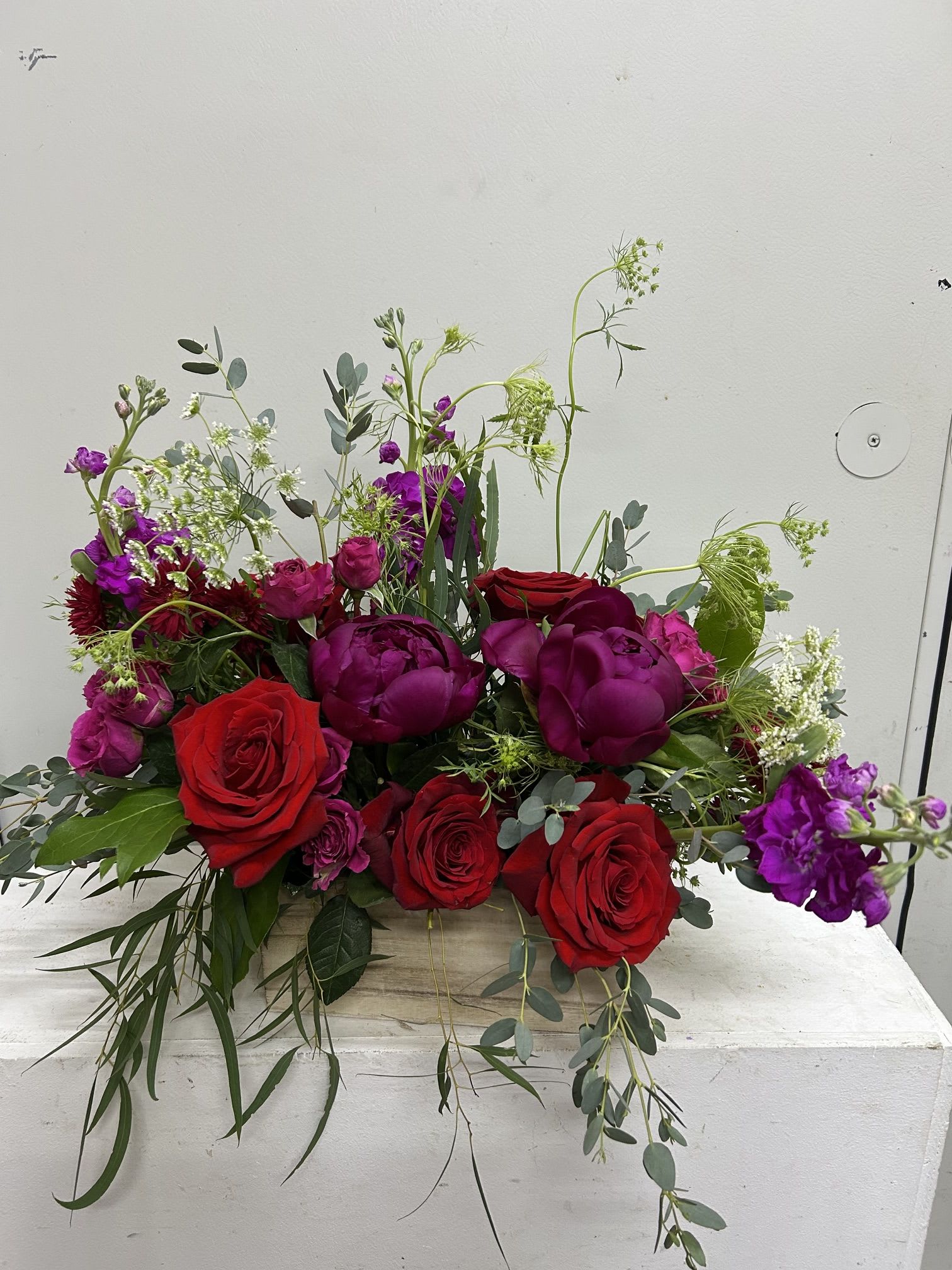 Red Sunshine - Saturated red roses and deep purple blooms create a bold, romantic statement in this low, garden-style flower arrangement-perfect as an anniversary bouquet, birthday surprise, or a spontaneous love-and-romance gesture. The mix of roses and spray roses with rich purples and pops of pink reads dramatic yet approachable, the kind of gift that makes the person you're thinking of feel vividly noticed.  Packed into a rustic wooden box, the design is lush and abundant but sits low for use as a centerpiece or a bedside surprise. Airy white filler flowers and silvery eucalyptus add cool contrast and a soft, loose texture while long trailing greenery gently softens the edges. Size and stem count vary by selection; arrangements arrive ready to display in the box-trim stems and refresh the water as needed to extend bloom life.