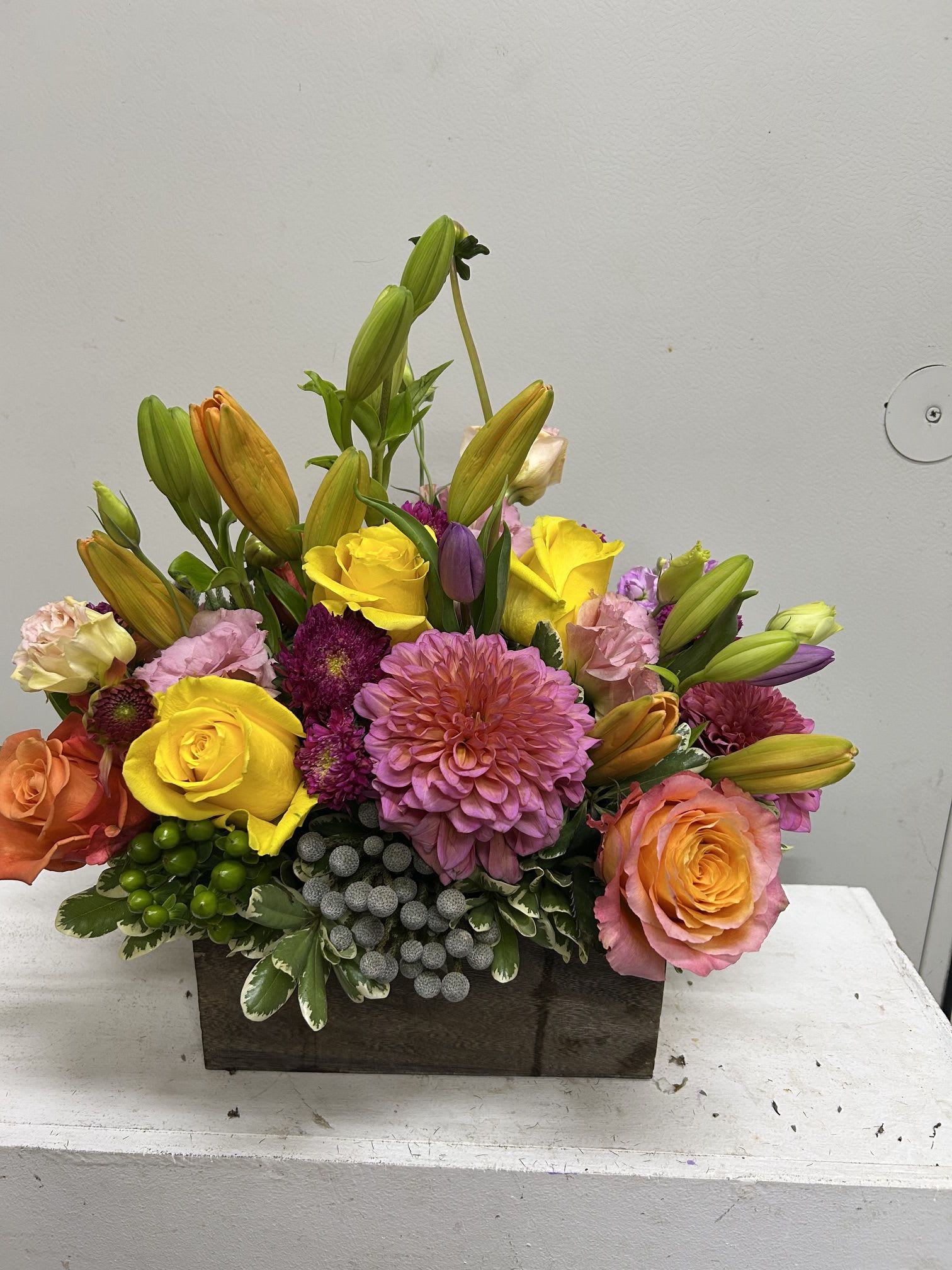 Spring Delight - A lively, garden-style centerpiece that brightens any birthday, congratulations, or thank-you moment-this low floral arrangement for a cheerful occasion places yellow roses and lily buds above a full carpet of dahlias, chrysanthemums, and lisianthus for instant impact. The mix of warm yellows, pinks and oranges with pops of purple gives the person you're thinking of a radiant, celebratory feeling the moment they see it.  Arranged densely in a rectangular wooden box, the design has a slightly formal finish while still feeling fresh and joyful. Yellow roses and a few upright lilies provide height and focal points, while dahlias, chrysanthemums and smaller lisianthus and accents create a textured, layered base; the compact shape makes it an ideal table centerpiece or desk brightener. This is the kind of bouquet that reads as thoughtful and festive-perfect for someone who deserves a sunny surprise.
