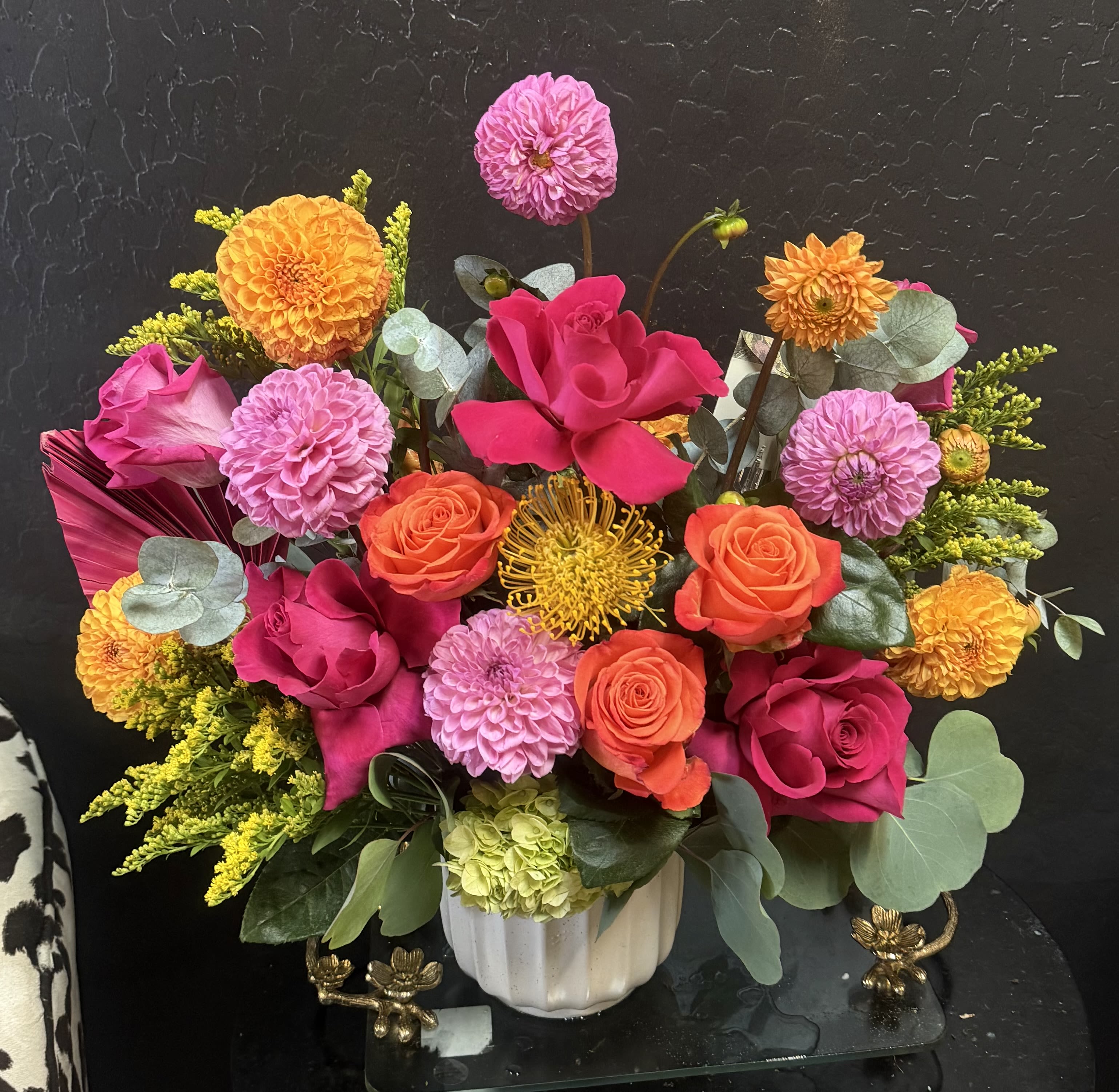 Tropical Dalhia Love Bouquet  - Shown in Premium Size Hot pink and orange roses take center stage in this Tropical Dahlia Love bouquet, a bold flower arrangement for birthdays, congratulations, or a cheerful "just because" surprise. The saturated mix of pink, orange, yellow, green, and purple creates an immediate, celebratory impression so the person you're thinking of feels truly seen the moment it arrives.  This dense, rounded mixed bouquet is presented in a white ceramic vase and layers hot pink and orange roses with dahlias, chrysanthemums, pale green hydrangea, daisies, and round magenta and lime-green blooms for rich, spherical texture. Yellow pincushion-style flowers and sprays of yellow filler mingle with eucalyptus-like foliage to add movement and bright contrast against the vivid petals, giving the design a polished, modern yet abundant feel.  Ideal for friends, family, or coworkers marking a happy moment, this arrangement works as a festive centerpiece or a bright thank-you gift that lifts the room. The bouquet is arranged to appear full in its ceramic vase; refresh the water every few days and trim stems as needed to help the blooms last longer. 