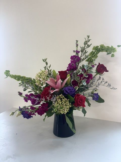 Prestige in Bloom - Prestige in Bloom is a striking display of modern luxury and vibrant beauty. Designed in a sleek black vase, this asymmetrical arrangement features premium blooms including rich red roses, fragrant hot pink stock, elegant purple lisianthus, graceful pink lilies, vibrant green hydrangea, and towering Bells of Ireland. Its bold colors and artistic design create a sophisticated statement that is perfect for celebrating life's most memorable moments.