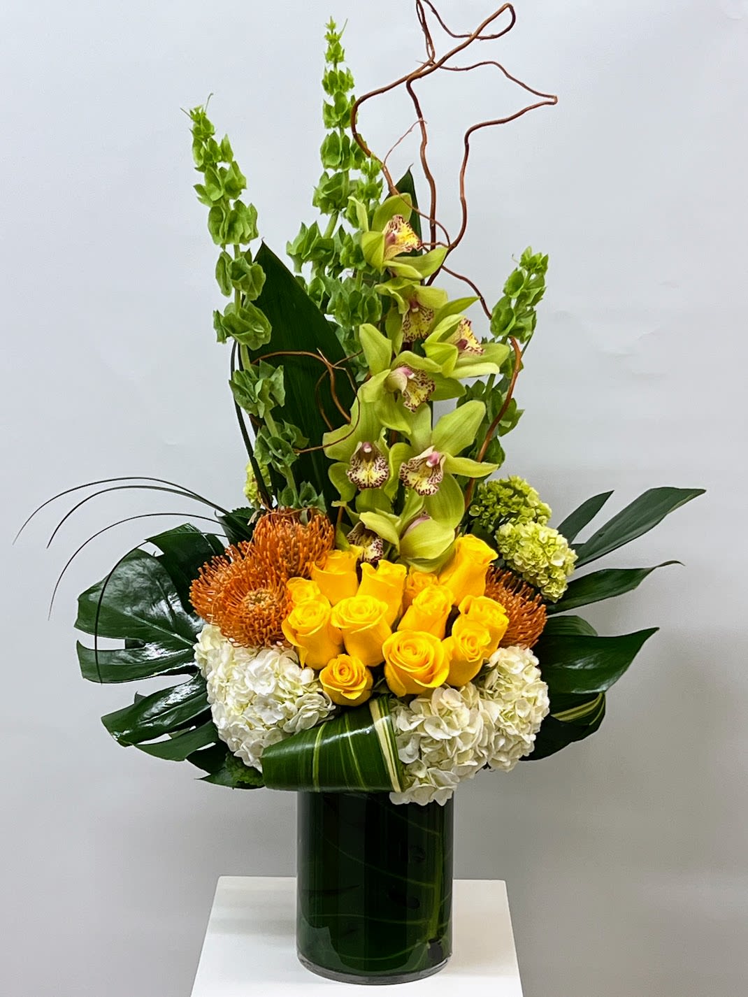 Tall floral arrangement with yellow roses, green orchids, and white hydrangeas in a glass vase