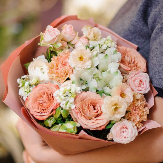 Handheld bouquet of peach, pink, and white flowers wrapped in pink paper