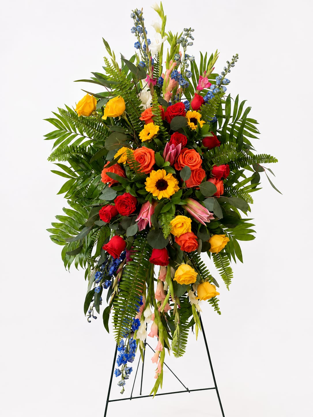 Sunlit Memories Standing Spray - Celebrate a life filled with love, joy, and treasured memories with the Sunlit Memories Standing Spray. This vibrant sympathy tribute is thoughtfully designed on an easel and features an uplifting blend of premium roses, cheerful sunflowers, elegant gladiolus, blue delphinium, and lush tropical greenery.  The warm palette of golden yellow, rich red, bright orange, soft pink, and calming blue creates a heartfelt expression of hope, gratitude, and remembrance. Its impressive vertical design makes it a meaningful focal point at funeral services, memorials, or celebrations of life, offering comfort to family and friends while honoring a loved one's legacy with beauty and grace.  Approximate dimensions: 42–48 inches tall and 28–32 inches wide (may vary slightly based on seasonal availability).