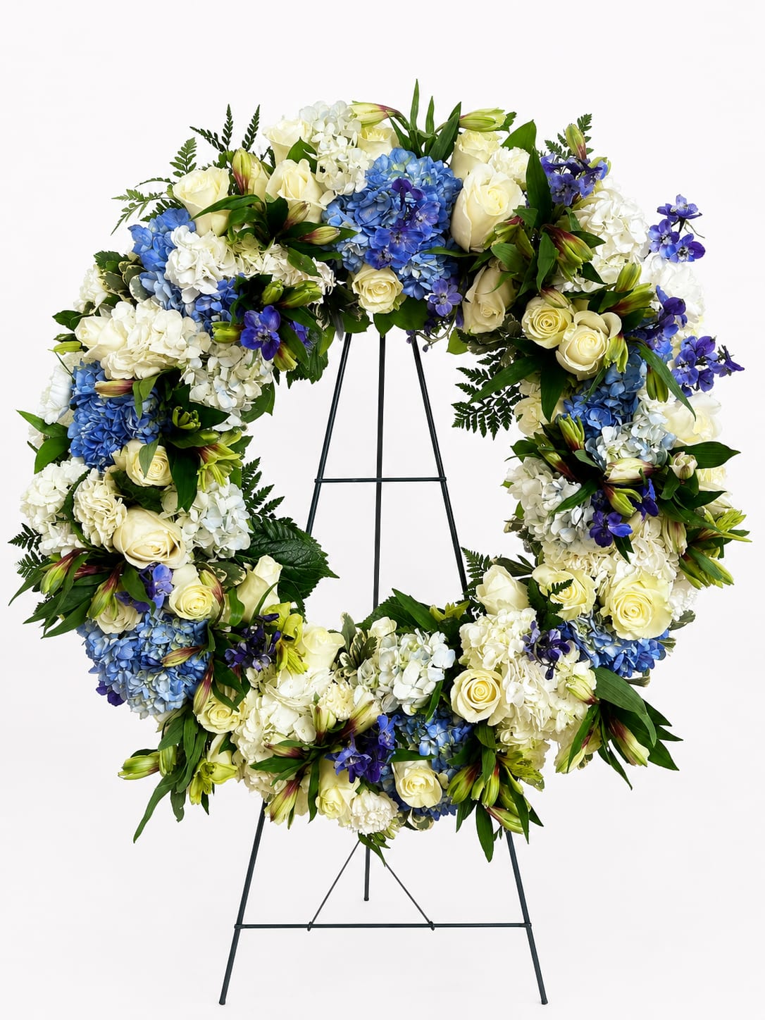 Serenity in Bloom Wreath - Honor a cherished life with the timeless beauty of the Serenity in Bloom Wreath. Designed as a symbol of eternal love and remembrance, this elegant standing wreath features a graceful blend of pristine white roses, lush white and blue hydrangeas, delicate lilies, and vibrant blue delphinium, accented with premium greenery for a naturally sophisticated finish.  The circular design represents everlasting love and the unbroken bond shared with those we hold dear. Its soothing palette of white and blue conveys peace, hope, and heartfelt sympathy, making it a meaningful tribute for funeral services, memorial ceremonies, or celebrations of life.  Handcrafted with care by expert florists and displayed on a standing easel, this wreath offers a beautiful expression of comfort and support during life's most difficult moments.  Key Features Premium white roses, hydrangeas, lilies, and blue delphinium Elegant circular wreath symbolizing eternal remembrance Accented with fresh seasonal greenery Displayed on a sturdy standing easel Suitable for funeral homes, churches, memorial services, and graveside ceremonies