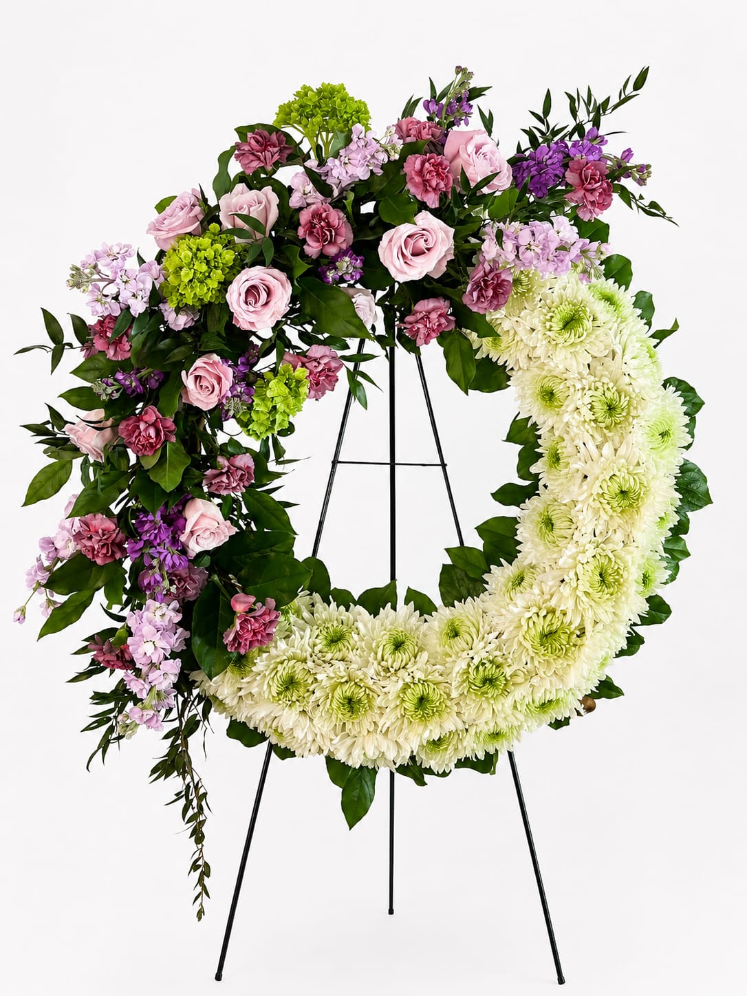 Graceful Devotion Wreath - Celebrate the memory of a loved one with the elegant beauty of the Graceful Devotion Wreath. This stunning half-and-half sympathy wreath combines a lush crescent of pristine white chrysanthemums with a romantic cascade of blush pink roses, mauve carnations, lavender stock, vibrant green hydrangeas, and fresh seasonal greenery.  Designed to symbolize everlasting love, peace, and cherished memories, the contrasting floral textures and soft color palette create a heartfelt tribute that offers comfort and hope during times of loss. Displayed on a standing easel, this handcrafted wreath is an exquisite choice for funeral services, memorial ceremonies, or celebrations of life.  Every arrangement is carefully created by skilled florists using premium blooms to ensure a beautiful and meaningful expression of sympathy.  Key Features Elegant mix of blush roses, mauve carnations, lavender stock, green hydrangeas, and white chrysanthemums Unique asymmetrical wreath design with modern garden-inspired styling Accented with lush premium greenery for added depth and movement Presented on a sturdy standing easel Ideal for funeral homes, churches, memorial services, and graveside tributes