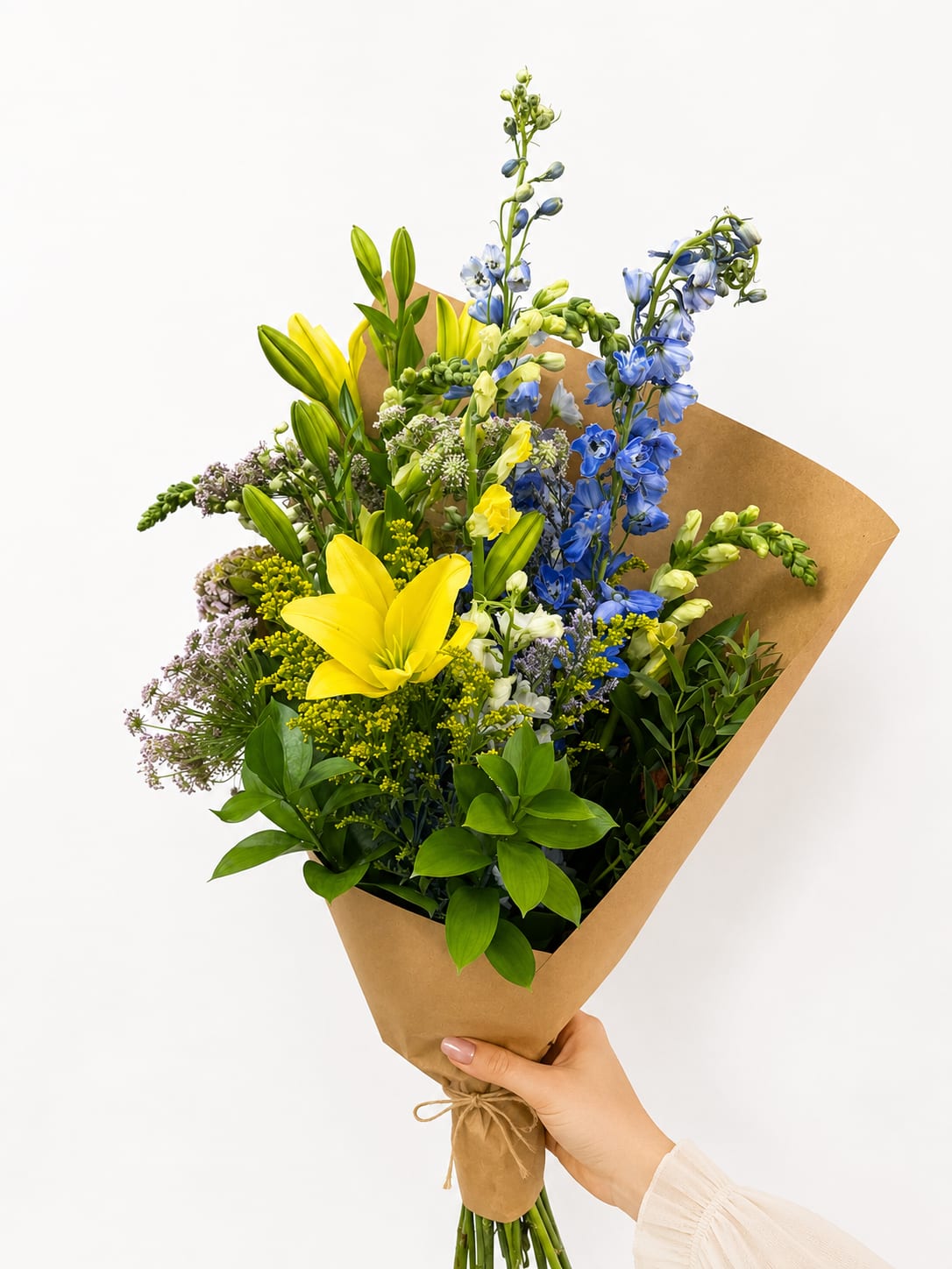 Golden Meadow Bliss Bouquet - Bright, cheerful, and naturally elegant, the Golden Meadow Bliss Bouquet is a hand-tied arrangement that celebrates the beauty of fresh seasonal blooms. Featuring radiant yellow lilies, striking blue delphinium, delicate white snapdragons, airy filler flowers, and lush premium greenery, this bouquet is wrapped in natural kraft paper for a modern garden-inspired presentation.  Perfect for birthdays, congratulations, get well wishes, thank-you gifts, or simply brightening someone's day, this bouquet offers a vibrant mix of color and texture that feels both sophisticated and uplifting. Handcrafted with fresh flowers and designed to impress, it makes a memorable gift for any occasion.