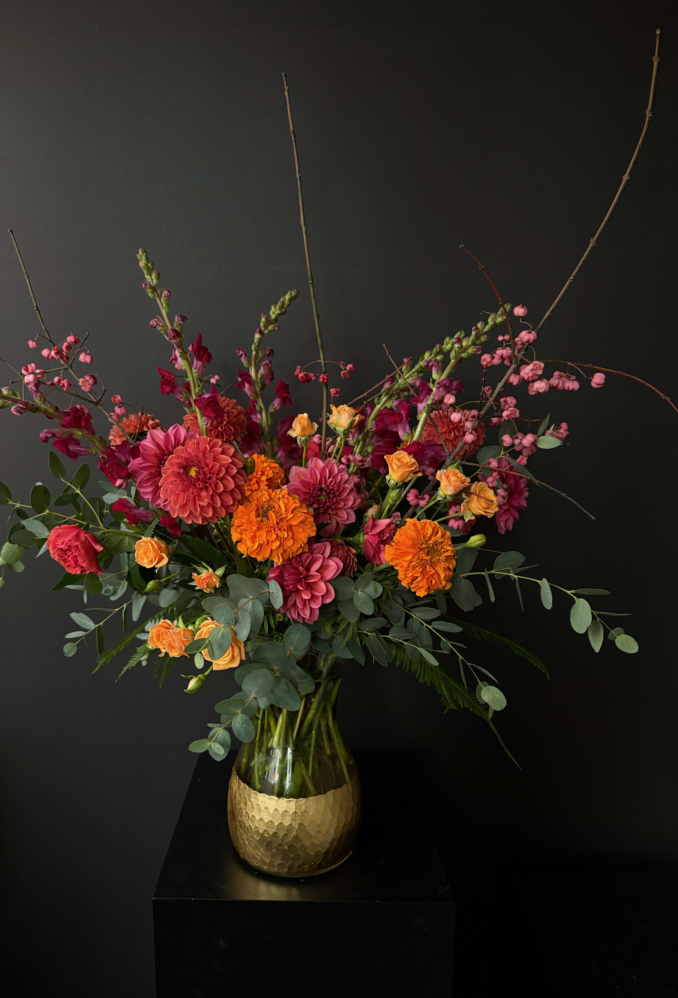 Tall arrangement of orange marigolds, pink dahlias, and spray roses in a glass vase with a gold base.