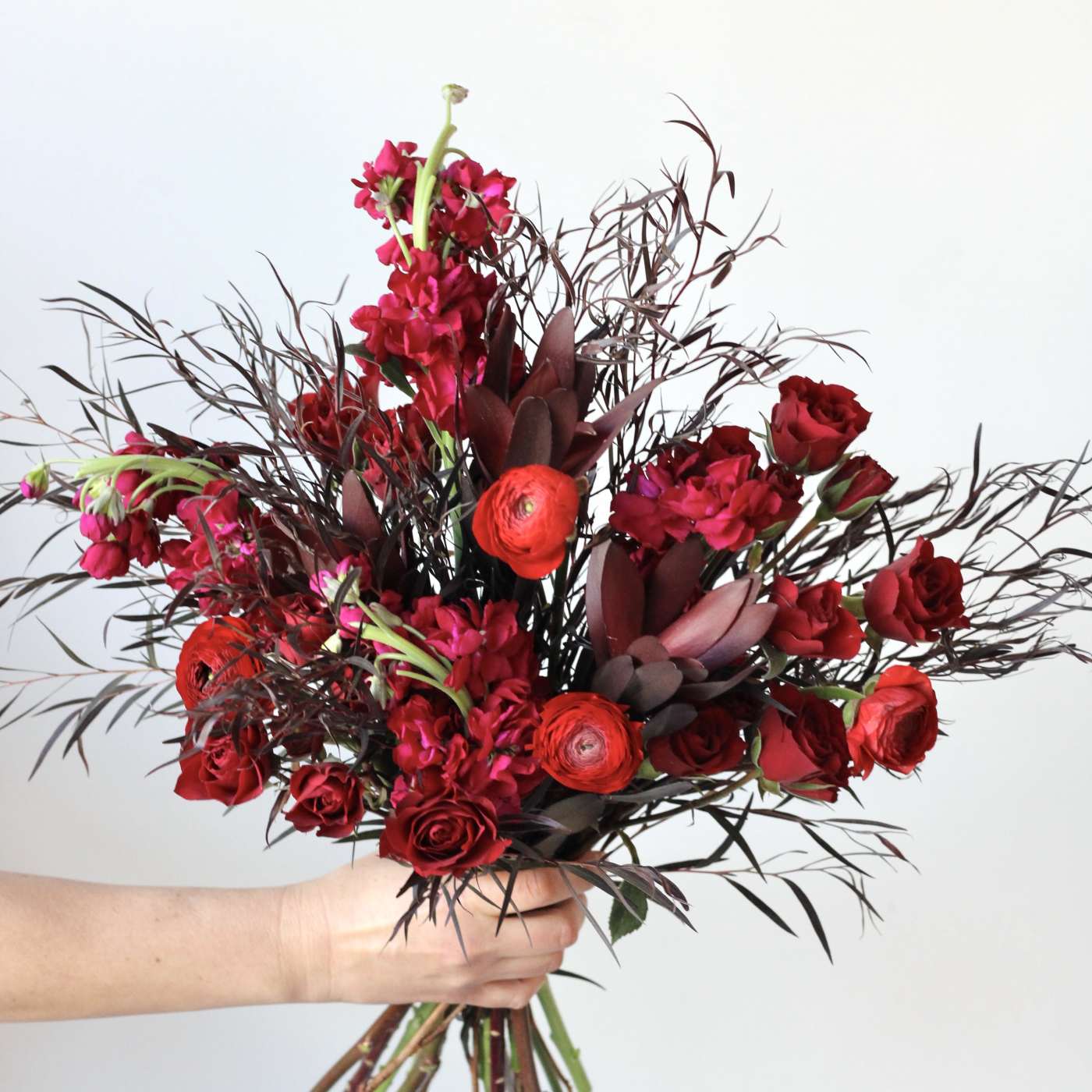 Handheld bouquet of red roses and red blooms with dark foliage