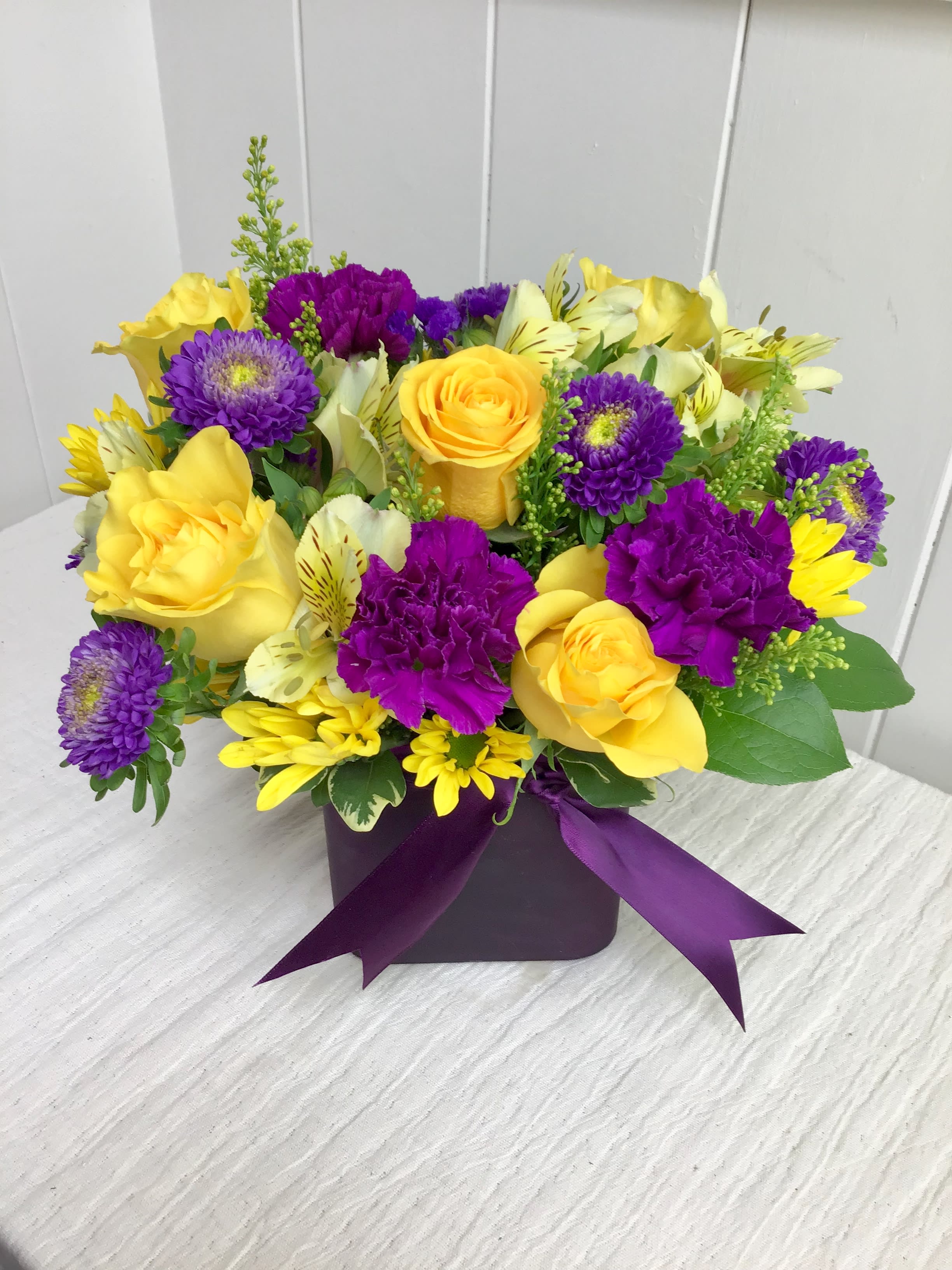 Low cube arrangement of yellow roses and daisies with purple carnations and asters in a purple vase with ribbon