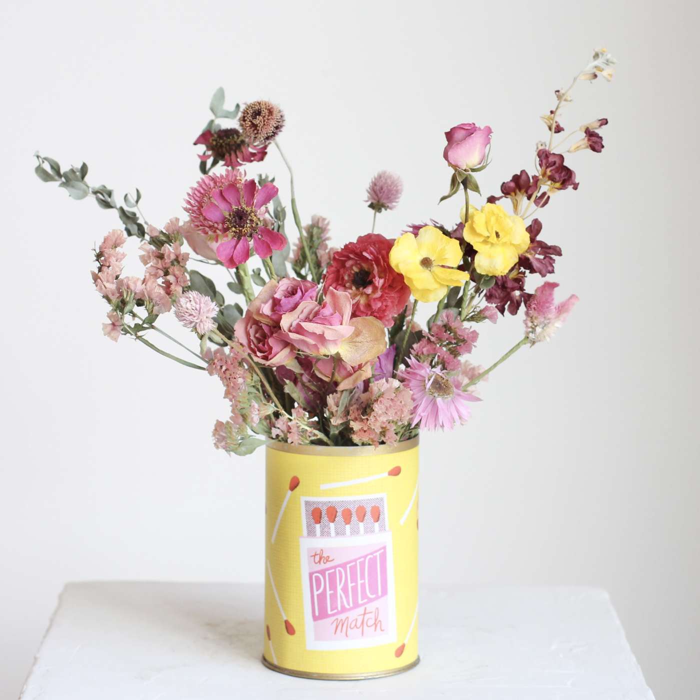 Mixed pink and yellow flowers in a yellow matchstick tin