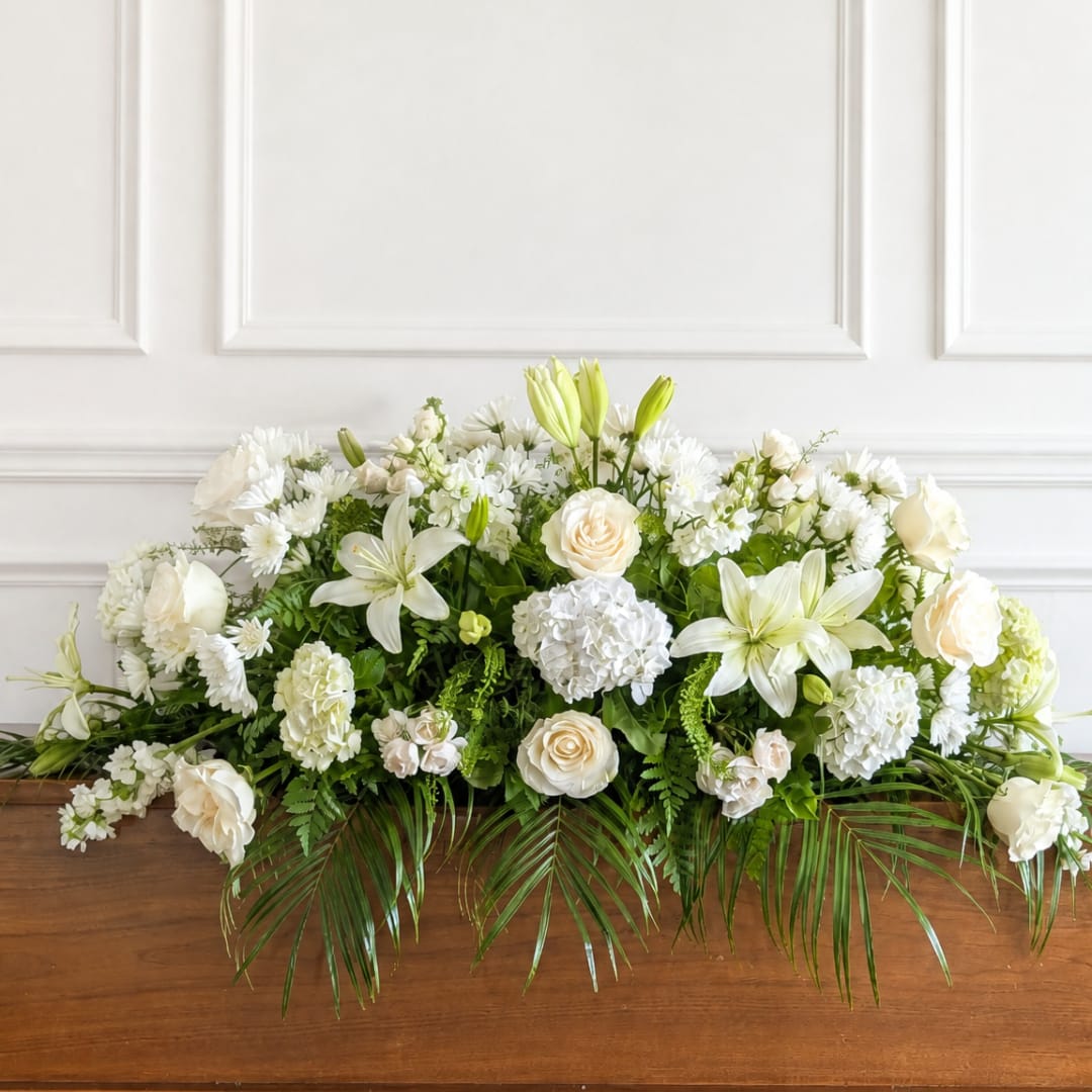 A Legacy in White - A Life Remembered is a traditional all-white casket spray created as a peaceful and heartfelt tribute to a life deeply loved. White and ivory roses, lilies, hydrangeas and complementary seasonal blooms are thoughtfully arranged among lush greenery to create a full, graceful presentation.  Designed in a classic casket spray style. Flower varieties may vary slightly based on availability while maintaining the all-white and ivory color palette and overall design.