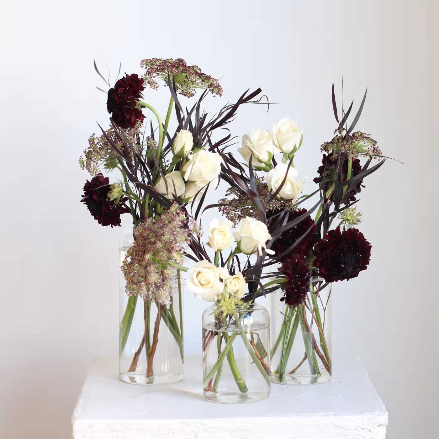 Three floral arrangements in clear glass vases with white roses and dark burgundy blooms.