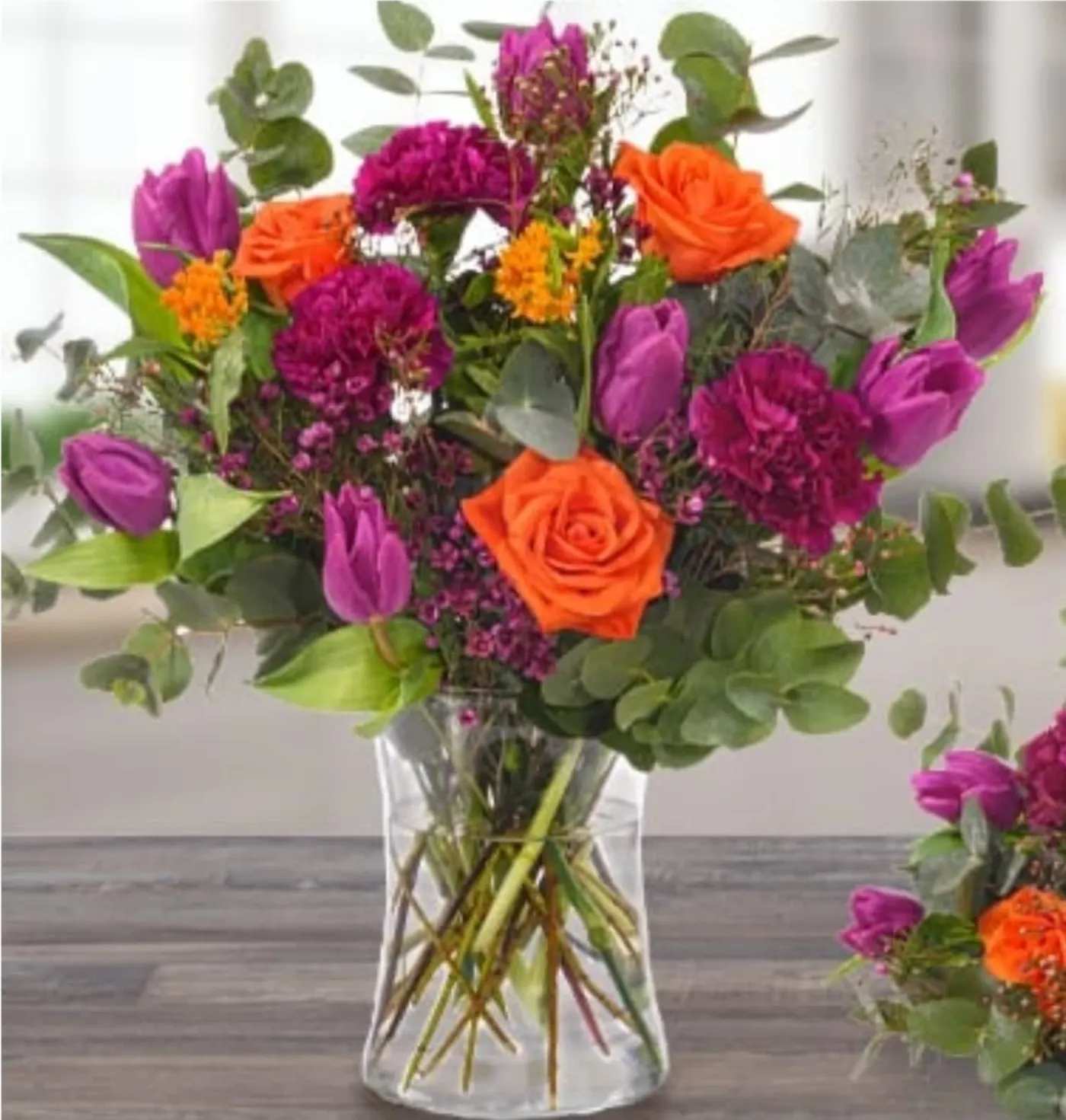 Bouquet of orange roses, purple tulips, and magenta flowers in a clear glass vase