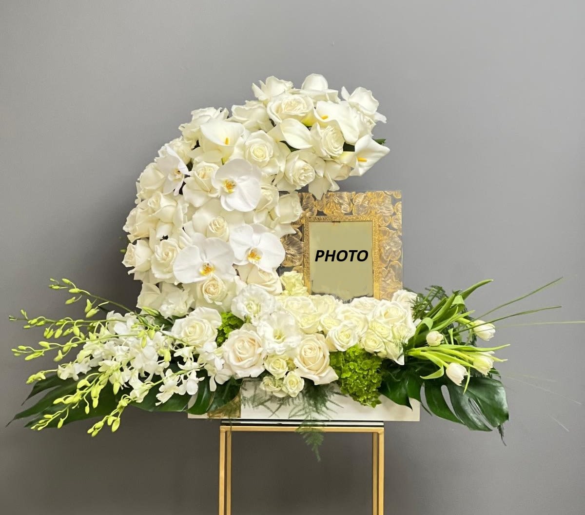 Eternal Grace Tribute - A serene and elegant sympathy arrangement designed to honor a life with grace and beauty. Featuring a harmonious blend of white roses, orchids, and seasonal blooms, this piece is thoughtfully composed around a framed photograph, creating a meaningful focal point for remembrance. Accented with soft greenery and presented in a refined structure with subtle gold elements, it offers a peaceful and heartfelt expression of love, respect, and lasting memory.