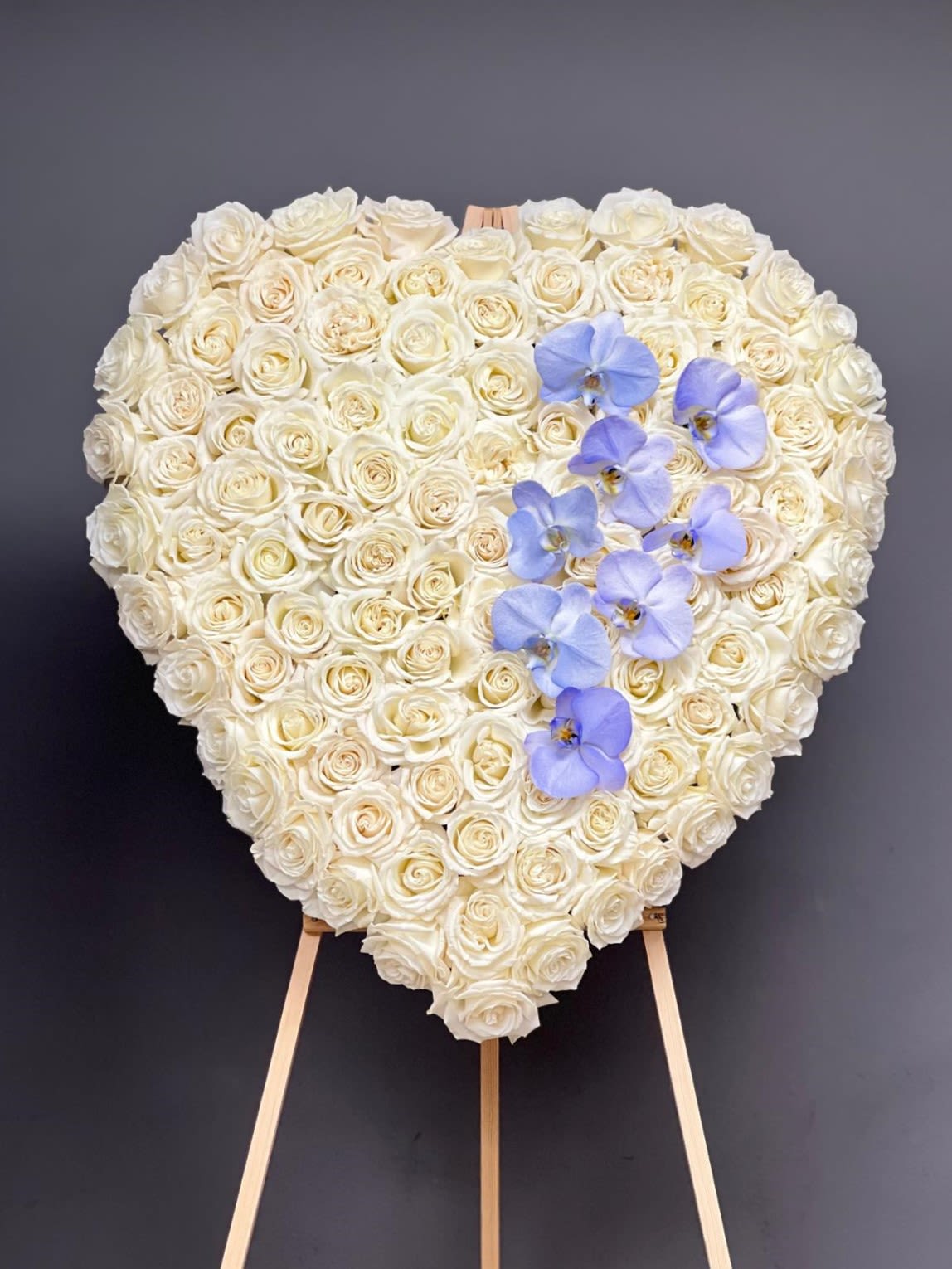 Eternal Love Rose Heart - A breathtaking heart-shaped standing tribute crafted with premium white roses and accented with delicate lavender orchids. Designed on an elegant easel, this arrangement expresses pure love, remembrance, and heartfelt sympathy with grace and sophistication. A printed ribbon or memorial card is included. Please indicate your preference in the “Special Instructions” section at checkout. They can be customized with your message that you can write in the "Card Message" section at checkout. Memorial cards are recommended for longer messages and personal notes while ribbons are best suited for short wording such as senders' (Family) names.