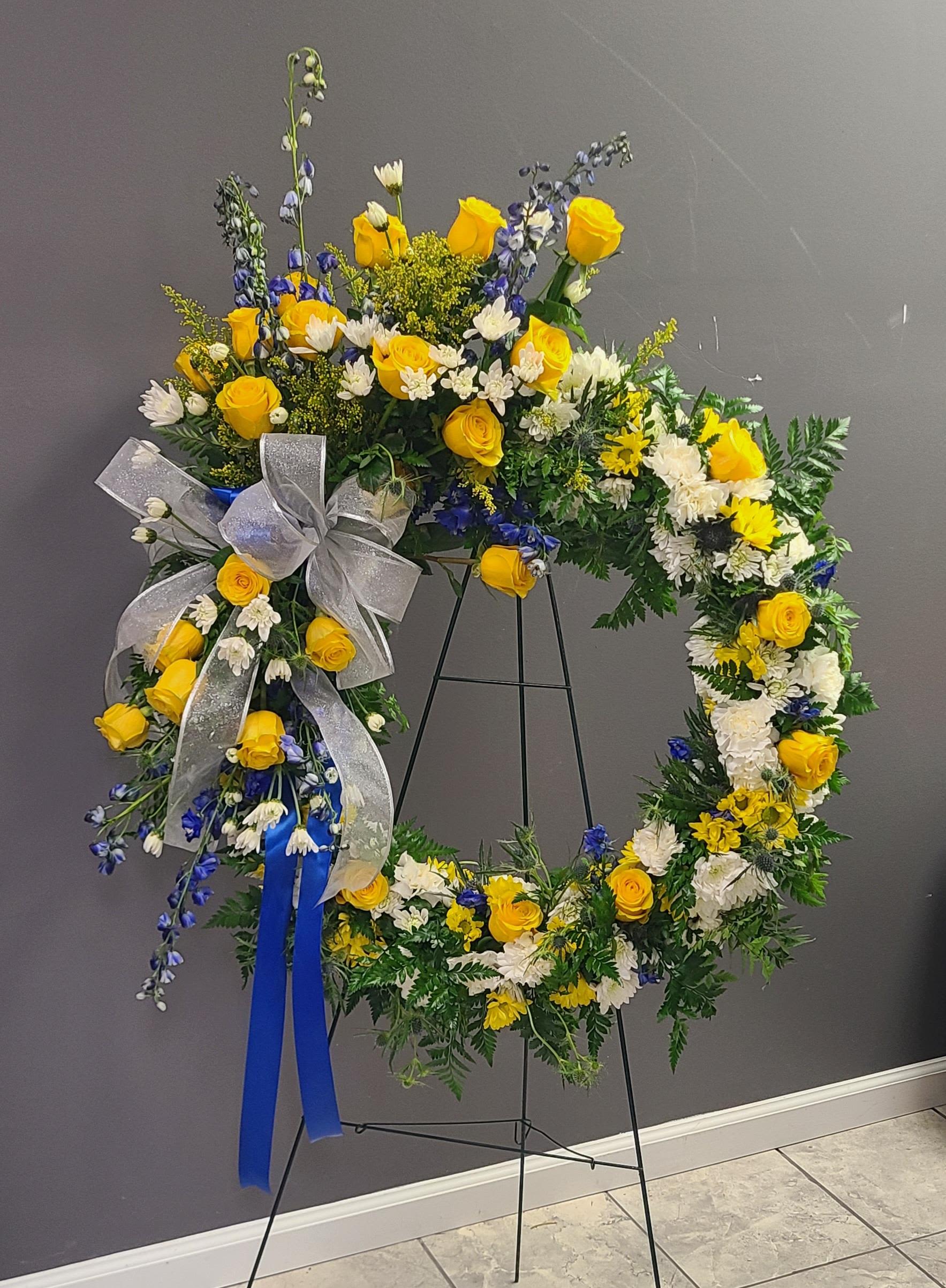 Standing Wreath for Family - Sunny Blue - A bright halo of yellow roses and white daisy-like blooms makes this standing wreath for family-Sunny Blue-feel both uplifting and respectfully reverent, perfect as a sympathy arrangement or to mark a memorial service. The circular design reads formal and ceremonial on the included black metal easel stand, while blue accent flowers and ribbons introduce a calm, hopeful note that speaks to remembrance and celebration of life.  The wreath is full and structured, combining vivid yellow roses with white daisies, snapdragons, and other blue-toned blooms arranged in a lush round shape. A wide silver bow and a long blue ribbon drape from the left, giving the piece a polished, traditional finish. Against the stand the colors-yellow, white, blue, and green foliage-create a clear, comforting contrast that will read beautifully in a chapel, reception, or graveside setting.  Made to honor a loved one, Sunny Blue offers a balance of brightness and dignity so the person you're remembering will feel genuinely celebrated. The arrangement arrives ready to display on the included black metal easel; for delivery or sizing questions, please contact us and we'll confirm availability and appropriate dimensions for your service.
