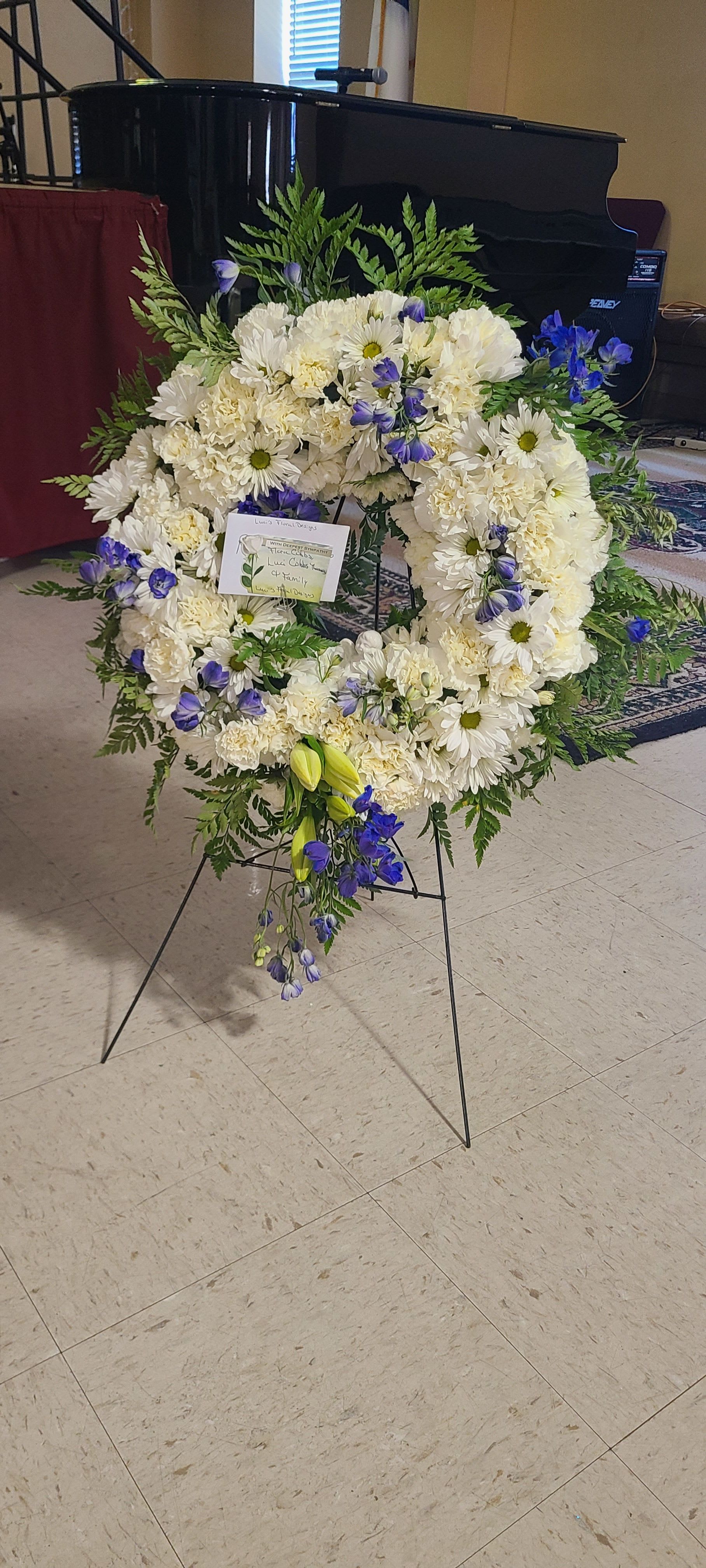 Standing Wreath Spray - A serene white sympathy wreath with vivid blue accents offers a respectful tribute for a funeral or memorial service. This standing wreath spray is designed for graveside services or a celebration of life, the kind of arrangement that helps the person you're honoring feel remembered and comforted.  Dense white chrysanthemums and clustered daisy-like blooms form the circular base, punctuated by vivid blue accent flowers and a few pale yellow lily buds for gentle contrast. Fern-like greenery frames the outer edge for a soft, layered silhouette. The arrangement arrives mounted on a black metal easel stand with a small card attached at the center, sized to make an elegant statement without overwhelming the service.  Appropriate for a formal memorial, this wreath sends a message of sympathy and quiet support while remaining tasteful and restrained. Place orders with recipient and service details in mind; care is simple - keep the stems hydrated until delivery and present on the easel at the service for best effect.