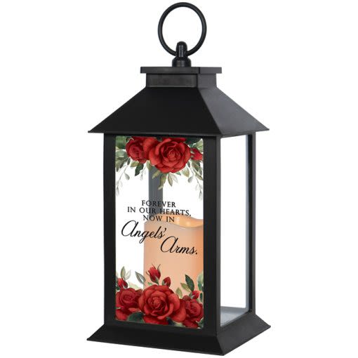 "Angels Arms" Lighted Sympathy Lantern - A black lantern framed in a classic rectangular silhouette offers a comforting memorial gift for sympathy and funeral occasions. The front glass panel is printed with rich red roses and a soft glowing candle, centered around the verse "Forever in our hearts, now in Angels' Arms," so the person you're honoring will feel remembered and gently celebrated whenever the lantern is displayed.  The lantern comes with a flameless LED candle (3 AAA batteries included) and a built-in automatic timer set to 6 hours on / 18 hours off, making it an easy, long-lasting keepsake for home or service displays. Weather-friendly materials and UV-printed glass preserve the printed roses and verse; the piece measures 5.5" wide by 13" high by 5.5" deep and can be hung by its loop handle or set free-standing.  For a more substantial tribute, choose the deluxe option to add a silk floral arrangement on the lid-an elegant way to personalize the lantern for the person you're thinking of.  Choose deluxe if you would like to add a Silk arrangement on lid of lantern.