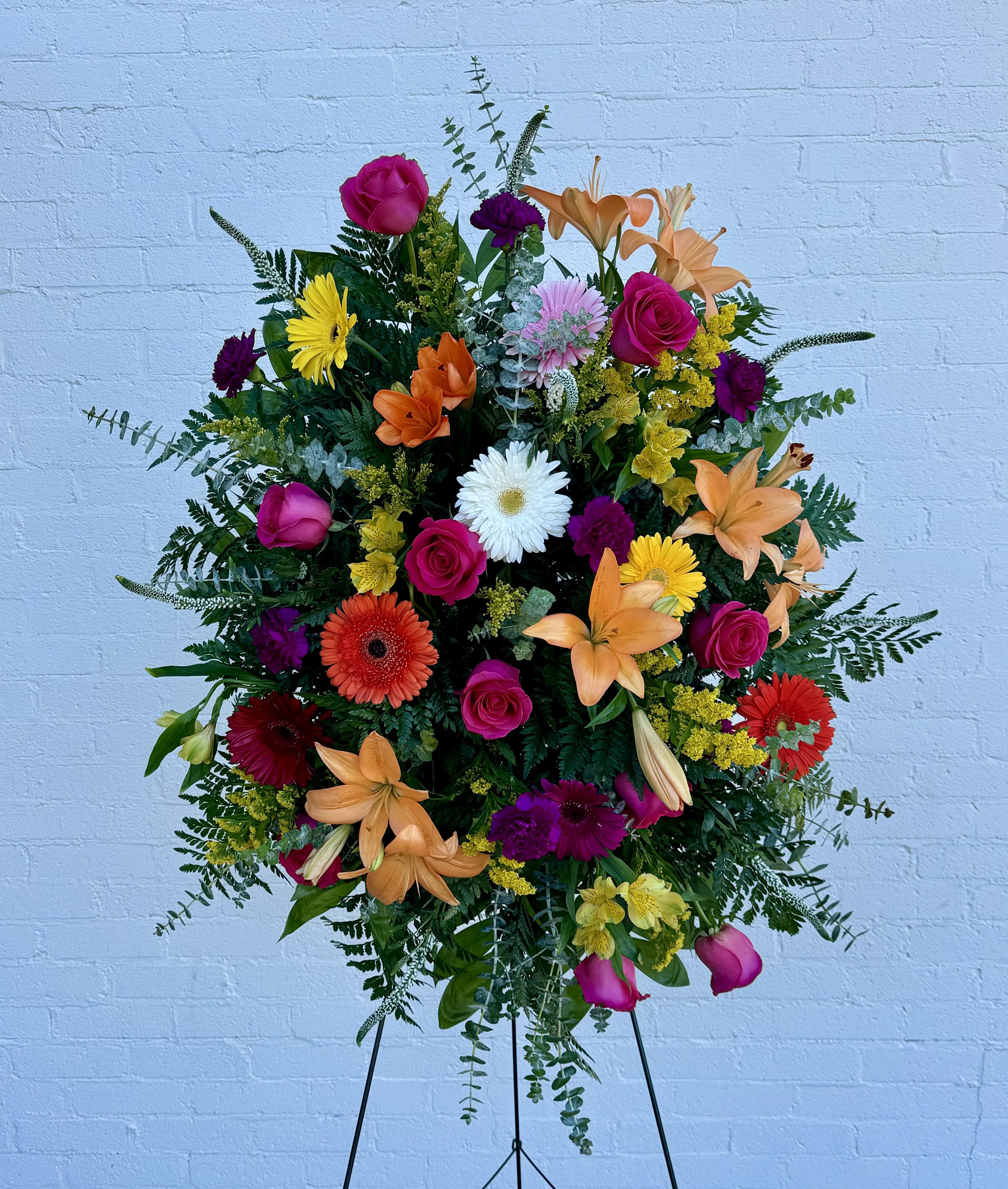 Colorful and Vivid Standing Spray - A dense, upright standing spray that commands attention with a vivid mix of hot pink roses, orange lilies and gerbera, purple carnations, white daisy-like blooms, and lush green foliage - perfect as a sympathy and funeral spray that celebrates a vibrant life. The layered, rounded silhouette presented on a standing easel reads formal and expressive, offering a bright, comforting presence at a funeral home or graveside while still feeling uplifting.  This arrangement includes alstroemeria, carnations, gerbera daisies, lilies, roses, and daisies in a high-contrast palette of purple, white, pink, orange, and mixed tones for a richly textured look. The design is full and abundant with a ceremonial scale that measures approximately 25" wide by 36" high and arrives ready on a standing easel. Our florists hand-design each piece, so exact blooms and color placement may vary with seasonal availability.