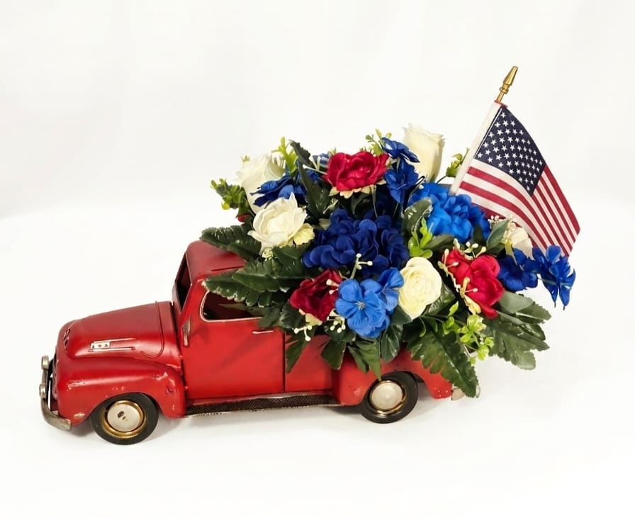 Patriotic Red, White &amp; Blue Pickup - A playful vintage-red toy truck overflowing with silk red, white &amp; blue blooms makes a cheerful patriotic flower arrangement perfect for Memorial Day, Fourth of July, Veterans Day, or any moment you want to celebrate a proud American spirit. This flower arrangement for seasonal decor and casual gifting places the occasion and recipient first-it's the kind of centerpiece that will make a veteran, neighbor, or host feel seen and appreciated.  Built into a small, decorative red pickup, the arrangement features silk daisies, carnations, tulips and mixed-color accents arranged in a dense, crafted cluster rising from the truck bed. The vivid reds, crisp whites and deep blues are softened by airy green foliage; the artificial blooms have layered petals for a tidy, long-lasting look that keeps its shape and color year after year. The whimsical toy truck adds instant personality and can be reused as tabletop decor.  Ideal for porch parties, patriotic table settings, or as a thoughtful gift to someone who loves Americana, this keepsake measures about 14" tall and 16" long-compact enough for tabletops yet bold enough to draw smiles. Because the flowers are silk, no watering or maintenance is required; simply dust occasionally to keep the colors bright.