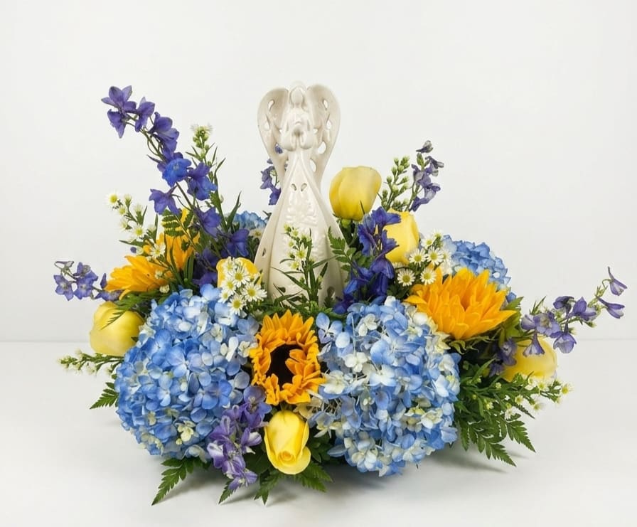 Ceramic Angel Floral Arrangement  - A serene, memorial-ready arrangement that still feels hopeful, centered around a white ceramic angel figurine-perfect as a sympathy arrangement or to mark a remembrance. The vertical presence of the angel gives this floral piece a devotional focal point while the bouquet's colors-soft blue, sunny yellow and white convey comfort and warmth to the person you're honoring.  Blue hydrangeas form a dense, cloud-like front while bright yellow roses and sunflower blooms bring uplifting contrast. Tall blue delphinium spikes rise around the angel, adding graceful height. The mix reads colorful yet calm, designed to feel supportive rather than flashy.  Choose this arrangement to express condolence or sympathy, the angel makes the sentiment clear while the vivid blooms celebrate a life. Sized to serve as a tabletop memorial or graveside tribute, it arrives ready to display. Would also make a great get well, birthday or just because gift. Angel is lighted by cord. 