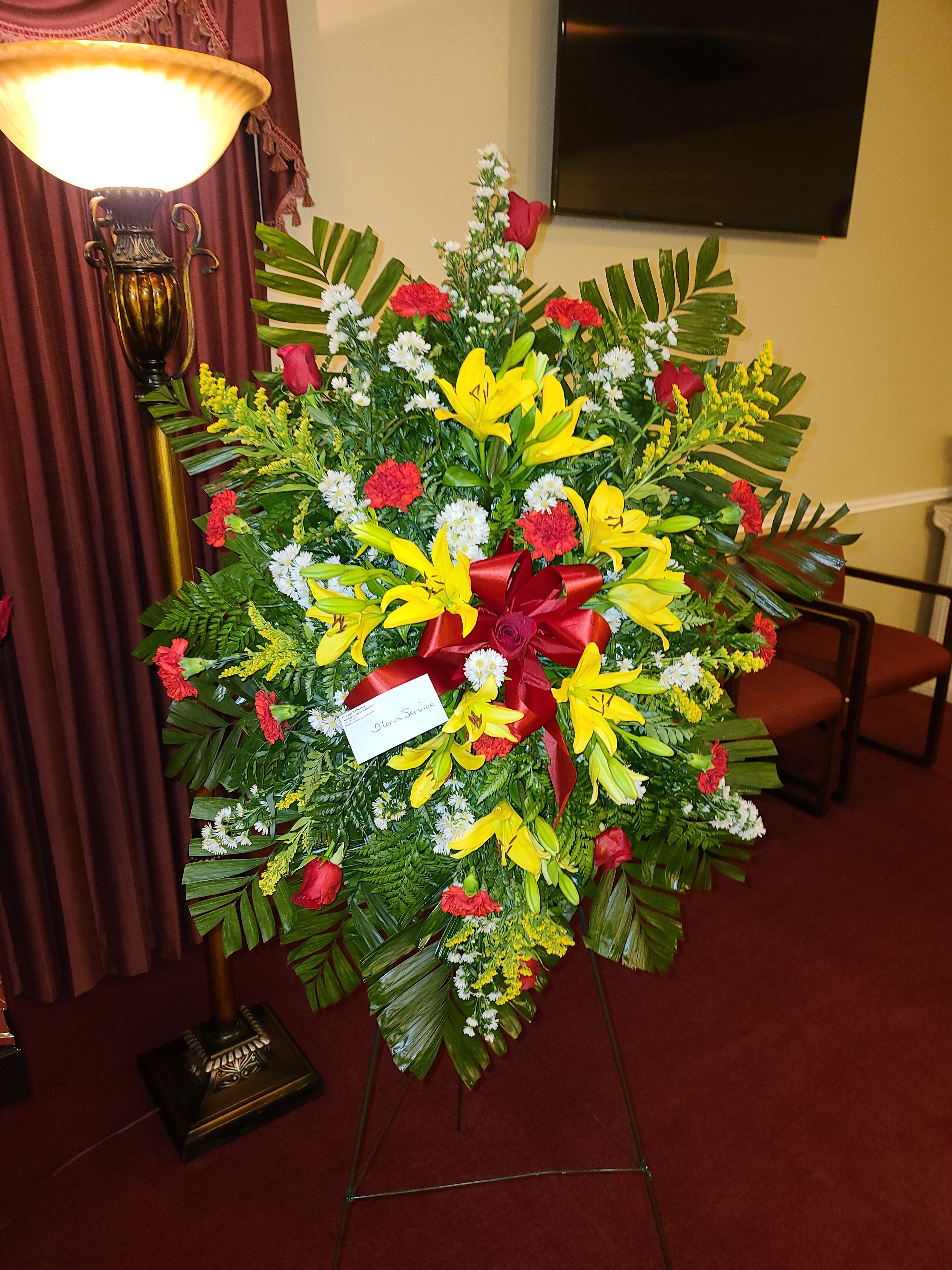 Large standing floral spray with yellow lilies, red roses, and a red ribbon