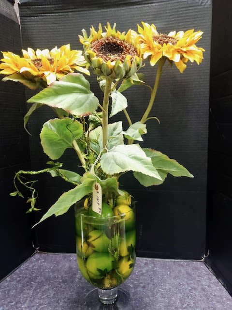 Three yellow sunflowers in a glass vase filled with green apples