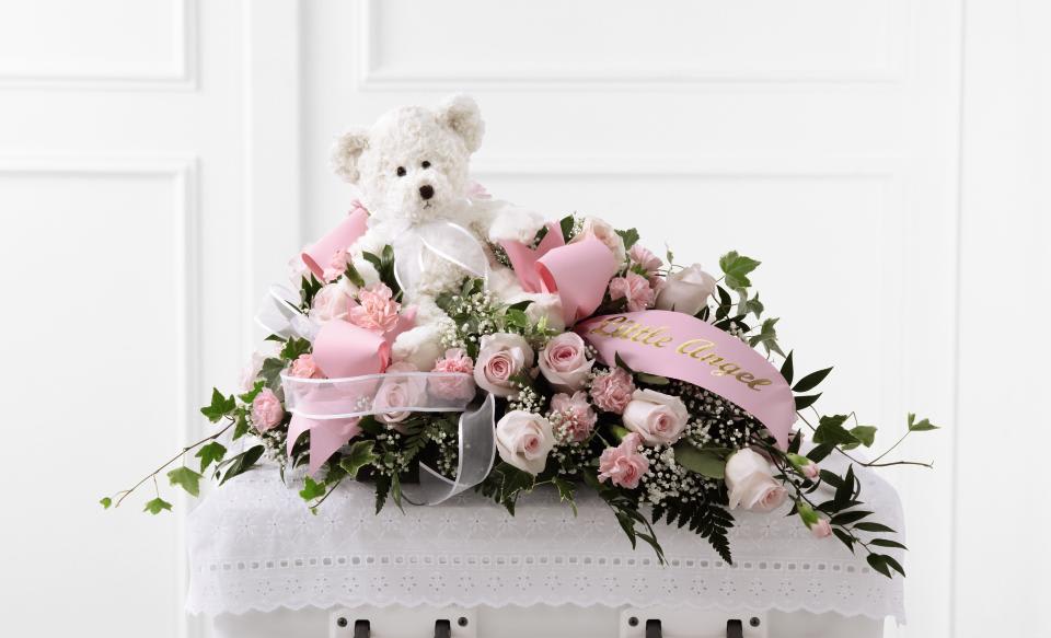 FTD Touch of Sympathy Casket Spray - The FTD Touch of Sympathy Casket Spray is a sweet and elegant  arrangement to honor a life taken too soon. Pale pink roses and pink  mini carnations are offset by baby's breath, ivy and lush greens to  create a beautiful arrangement intended to bedeck the top of the casket.  Accented with an ivory plush bear, a sheer white ribbon and a pink  ribbon that reads, ""Little Angel"" in gold metallic lettering, this  casket spray offers a lovely way to say your final farewell. 14""h x 24""w