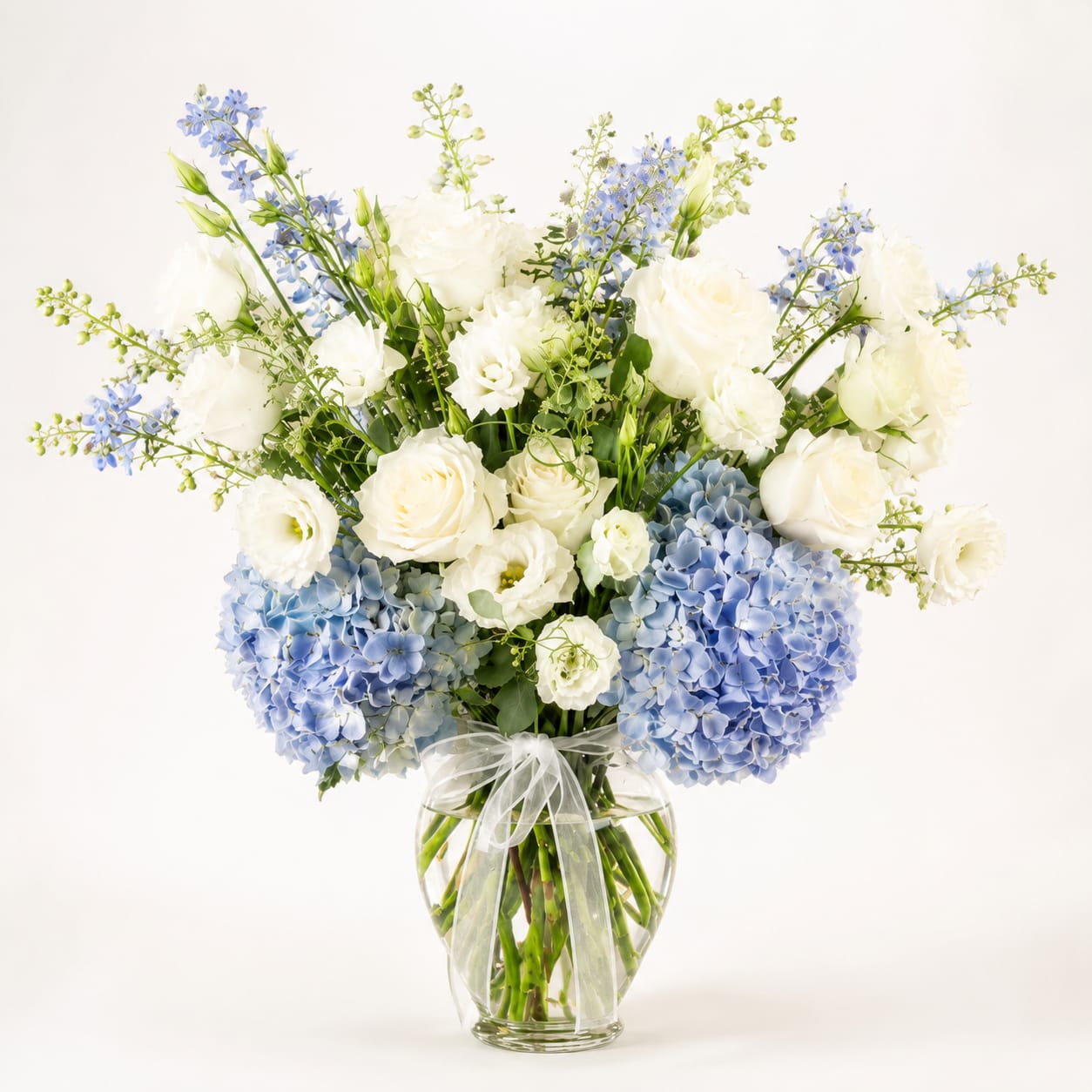 Blue Elegance - This graceful vase arrangement exudes serenity and sophistication with its soft palette of creamy white roses and blue hydrangeas. Accented with gentle greenery. Each bloom is carefully styled to evoke calm, purity, and tenderness. 