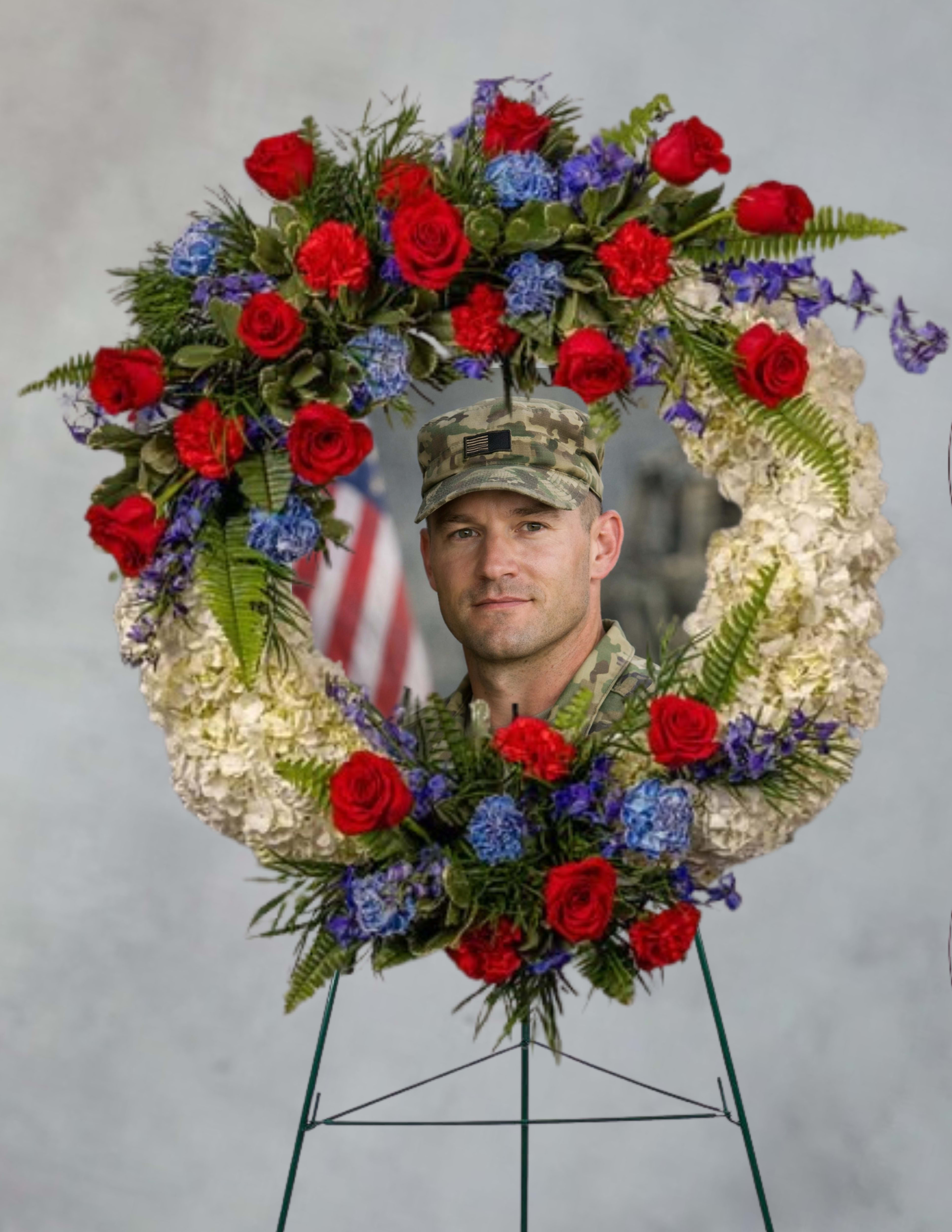 A hero’s grace  - A large floral wreath crafted for moments of honor and remembrance, perfect as a memorial tribute or commemoration display. This flower arrangement for a funeral or memorial service presents a patriotic red, white, and blue palette that quietly conveys respect and gratitude-an appropriate choice for Veterans Day, a military funeral, or to mark an anniversary of a loved one's passing.  Densely composed on a sturdy metal wreath stand, the design pairs bright red roses with clusters of blue blooms and a broad band of white flowers to form the heart motif. Fern-like green foliage frames and fills the shape, adding texture and a formal, structured silhouette. The arrangement reads ceremonial from a distance yet rewards closer viewing with the contrast of smooth rose petals against softer hydrangea-like clusters and leafy accents.  For the person being honored, this piece communicates heartfelt reverence and patriotic sentiment; it's the kind of tribute that helps family and friends feel their loved one is remembered with dignity. Mounted and ready to display, the wreath is sized for prominent placement at a service; minimal care is needed-keep shaded and mist sparingly if storing before the event to preserve freshness.