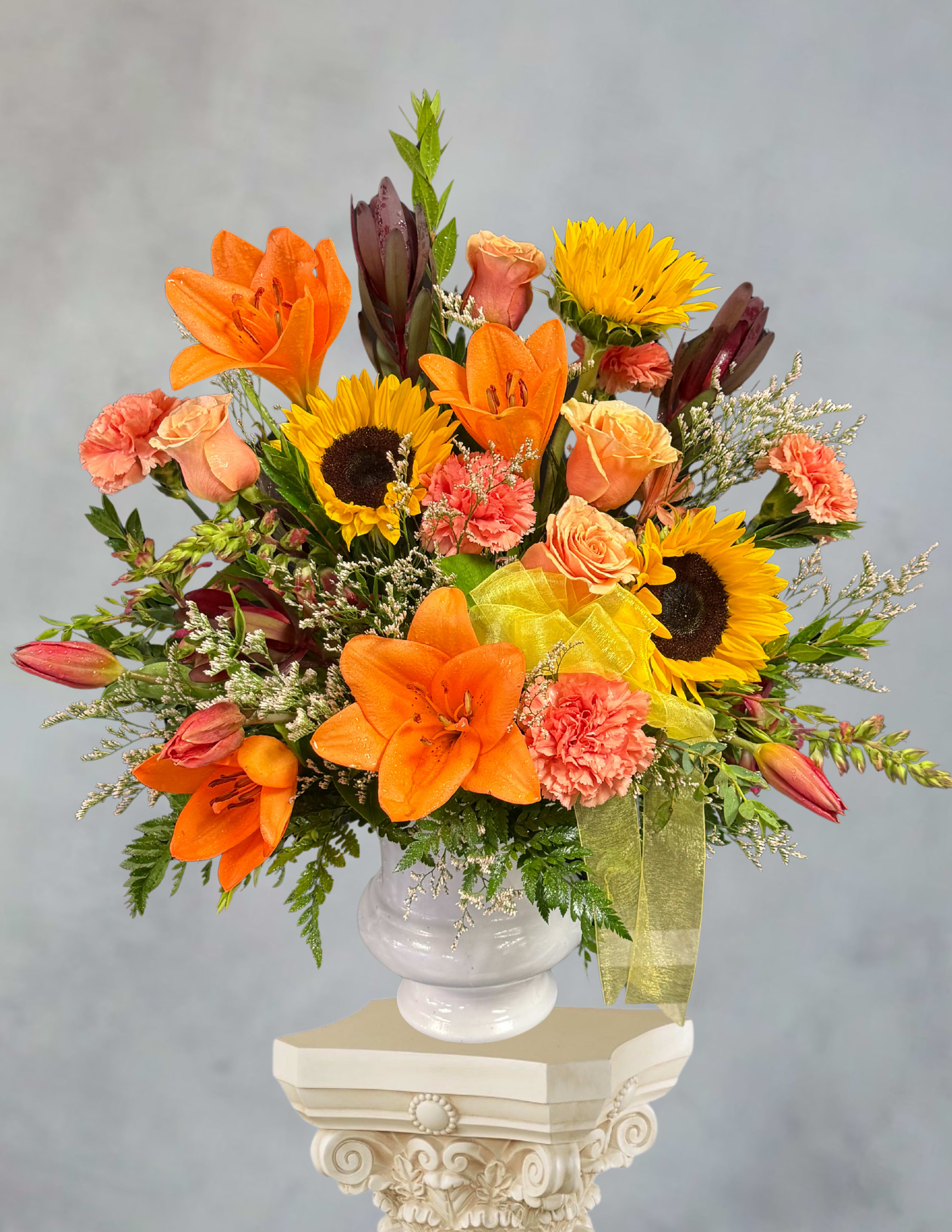 Golden pathway - Bright orange lilies and sunny sunflowers create an immediate sense of warmth and uplift-the kind of flower arrangement that feels like a golden pathway for someone who needs comfort or celebration. As a sympathy or funeral arrangement this flower arrangement reads as a gentle, hopeful tribute.  This full, layered design pairs peach roses and coral carnations with airy filler for texture, all arranged at varying heights in a rounded white ceramic vase tied with a yellow ribbon. The palette of orange, yellow, pink and white balances bold color with soft accents: the lilies and sunflowers provide visual impact, the roses add tenderness, and the carnations give lasting volume. The white vase keeps the look clean and timeless.  Thoughtfully composed to convey warmth without excess, this arrangement suits bedside tables, memorial services, or any room that needs a bright, reassuring presence. Vase dimensions and exact stem counts are available at checkout-select delivery timing to ensure the flowers arrive fresh when the moment matters most.